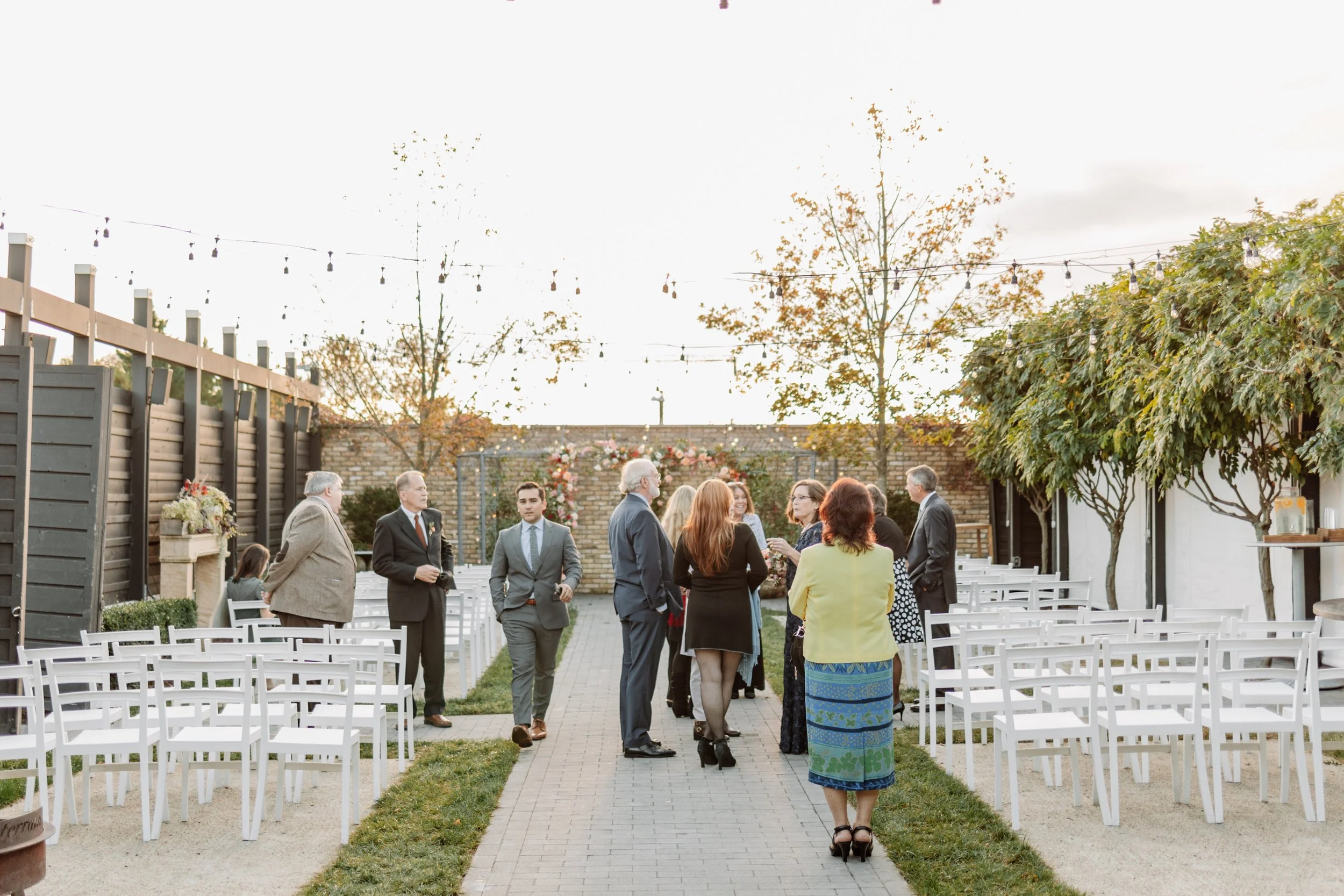 Guests mingling in an outdoor garden wedding setting with white chairs, string lights, and autumn trees