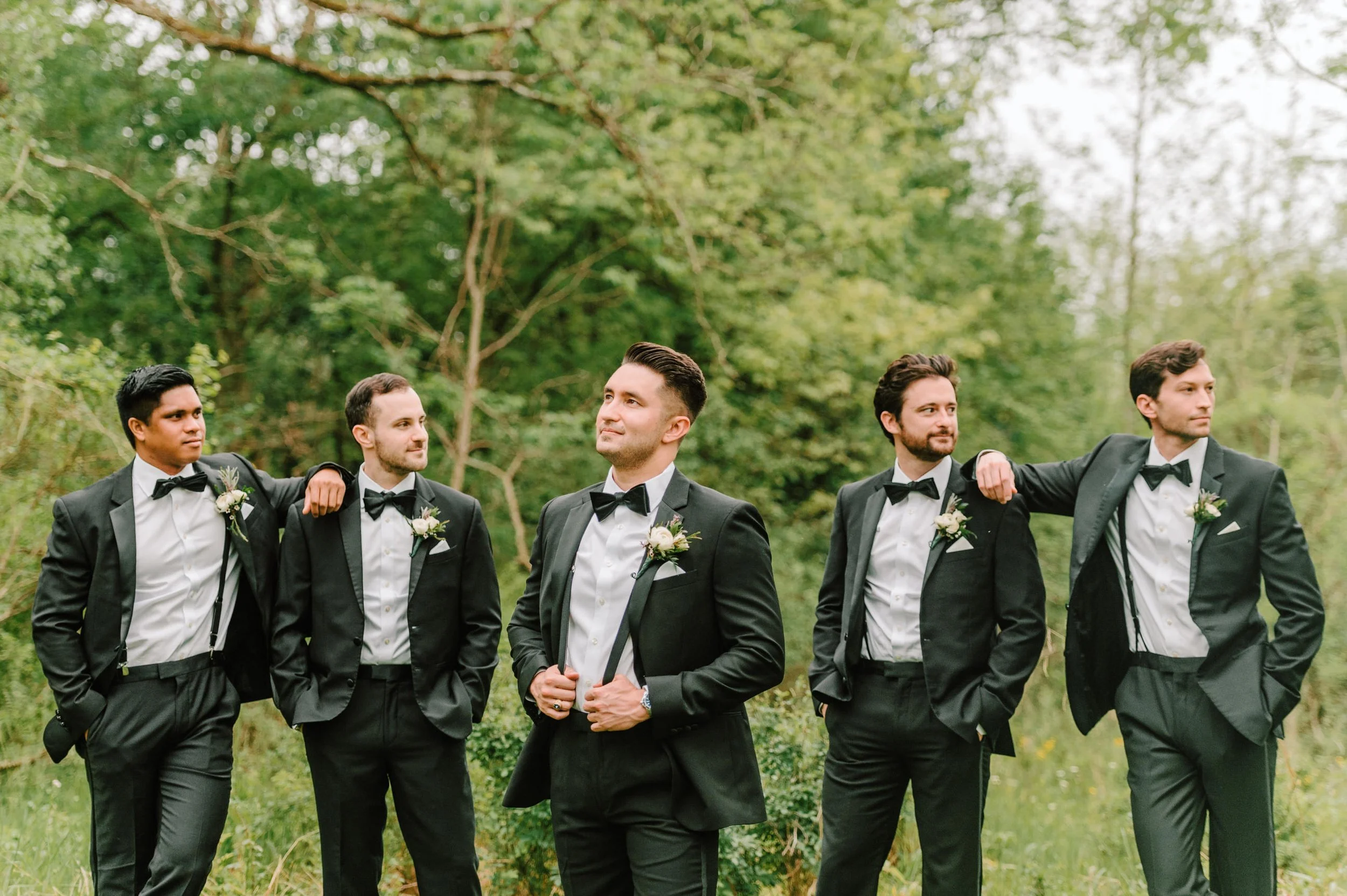 Group of five men in tuxedos and bow ties standing outdoors in a forest, with two men on each end resting their arms on the men's shoulders in the center.