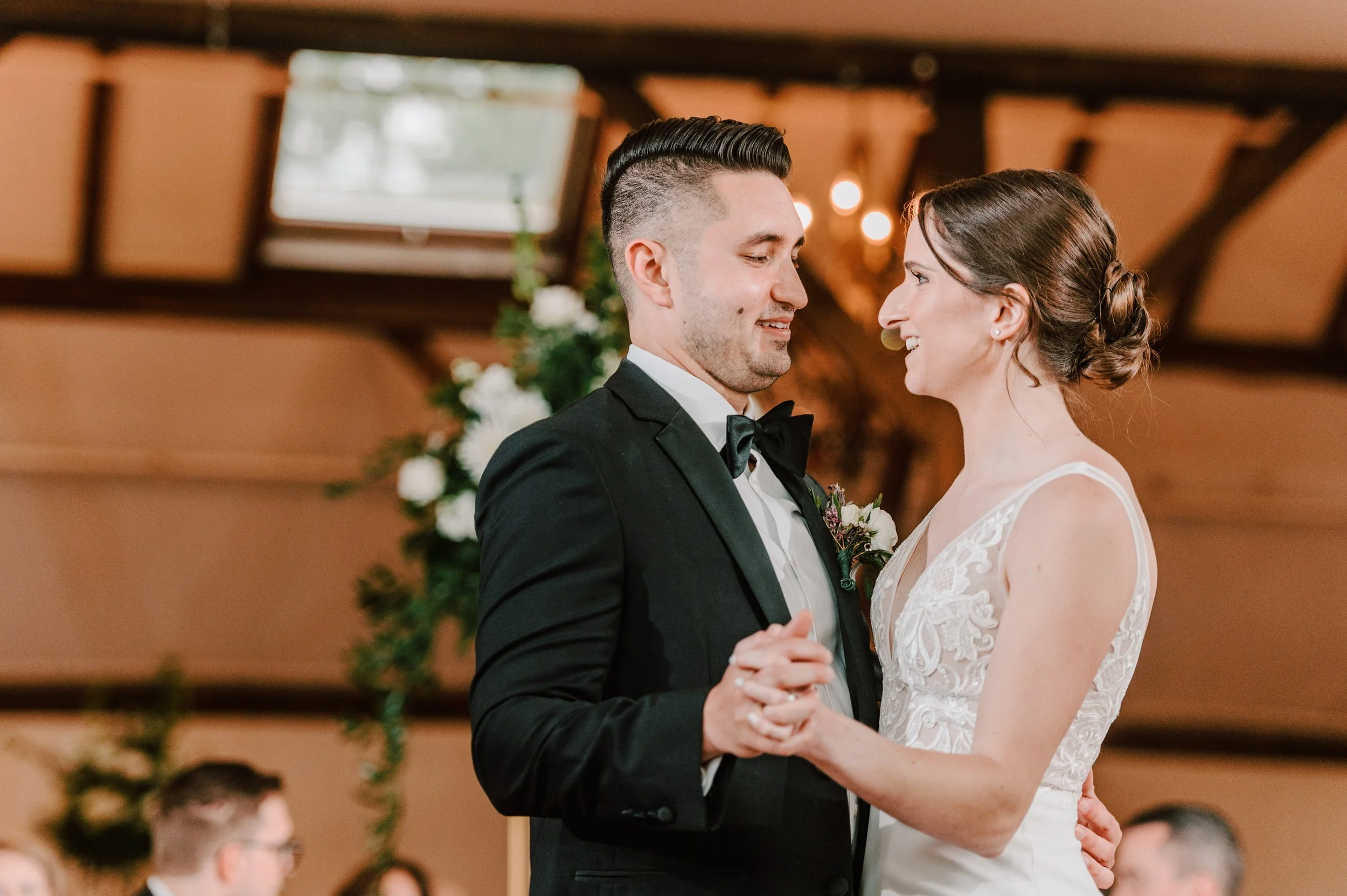 A newlywed couple, dressed in wedding attire, sharing a dance at their wedding reception indoors with guests seated at tables in the background.