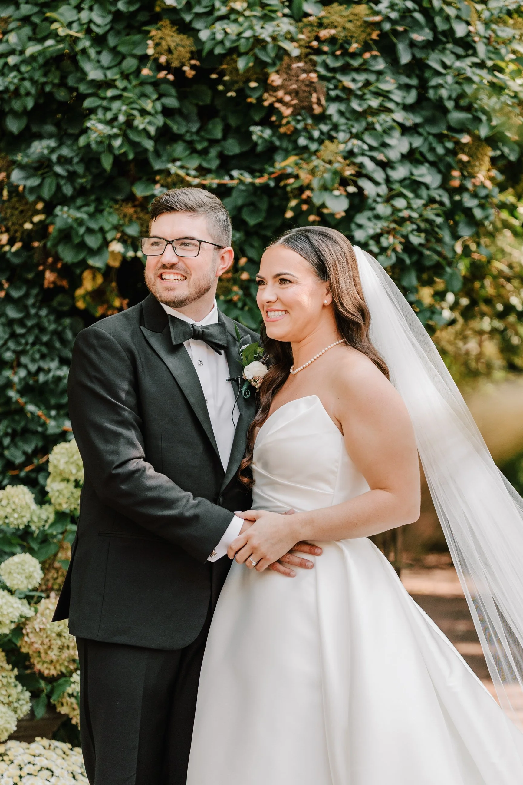 Bride and groom in wedding attire standing outdoors with greenery in the background.