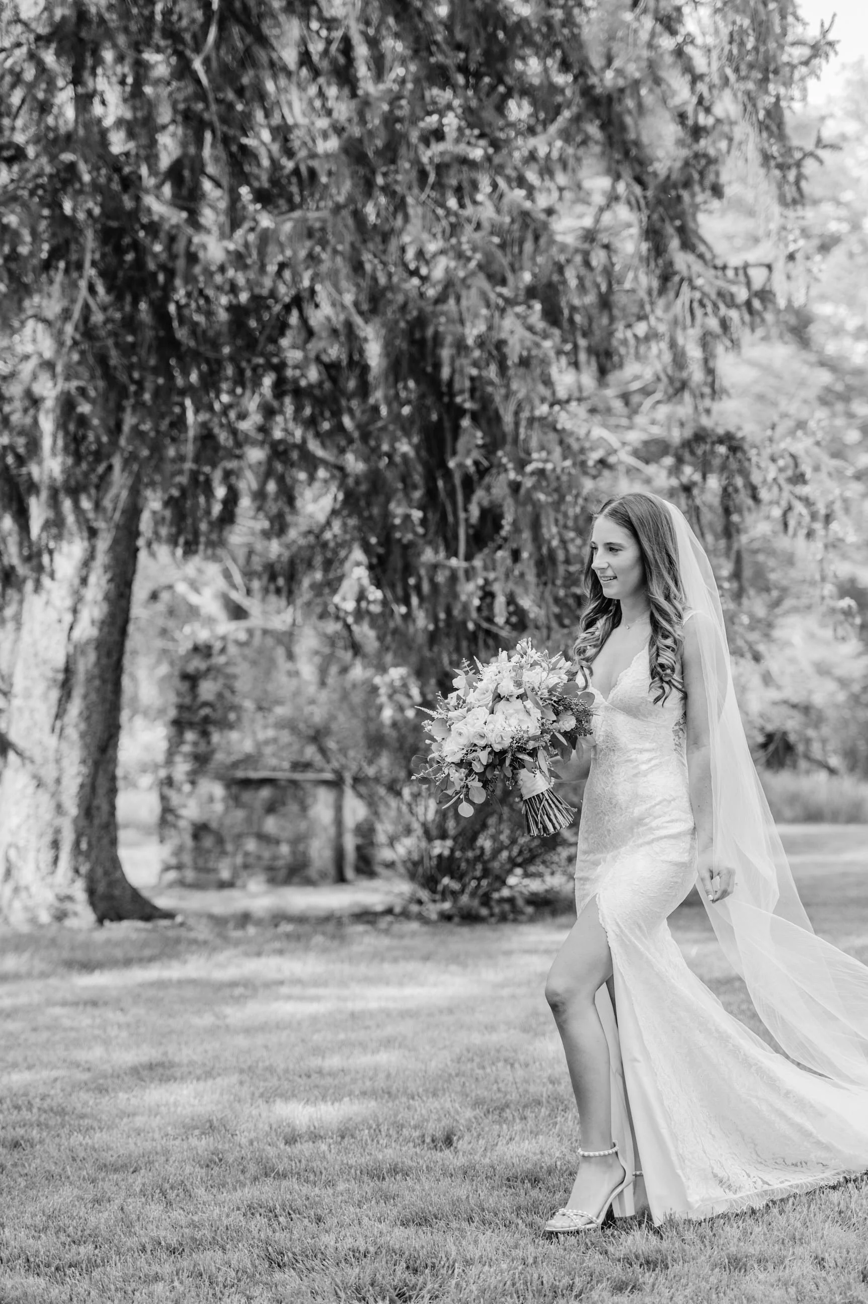 A bride with long hair wearing a lace wedding dress and veil, holding a large bouquet of flowers, standing on grass outdoors with trees in the background.