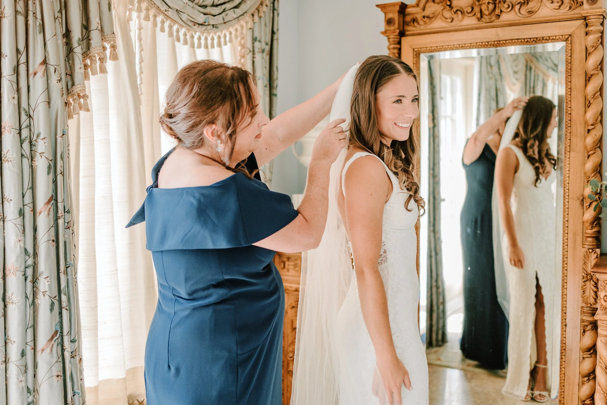 A woman in a navy dress helps a bride put on her veil in front of a mirror in a room with floral curtains and a large wooden-framed mirror.