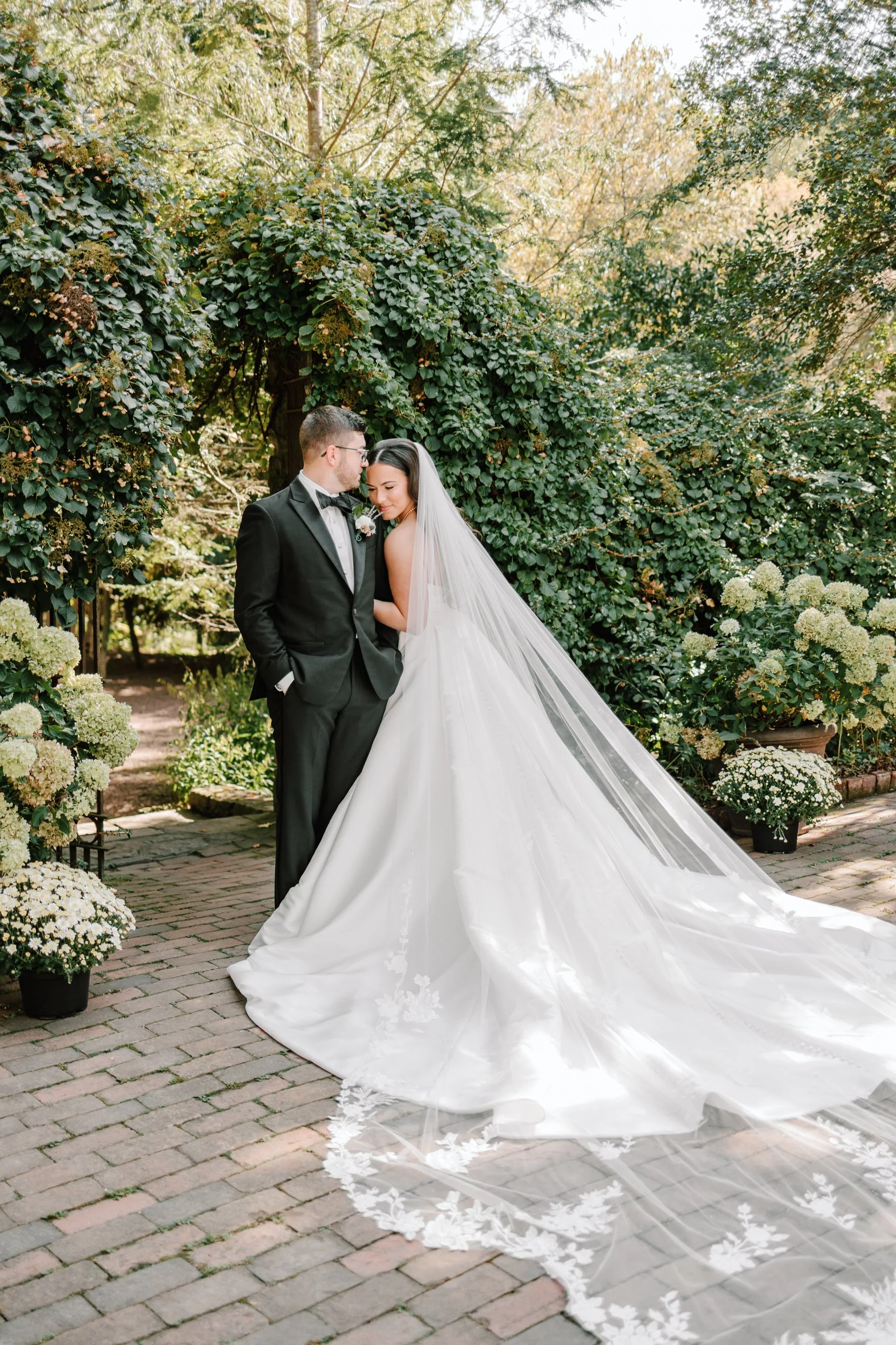 A bride and groom standing close together in a garden, with the bride wearing a white wedding gown and long veil, and the groom in a black tuxedo. The surrounding area has lush greenery and potted white flowers.