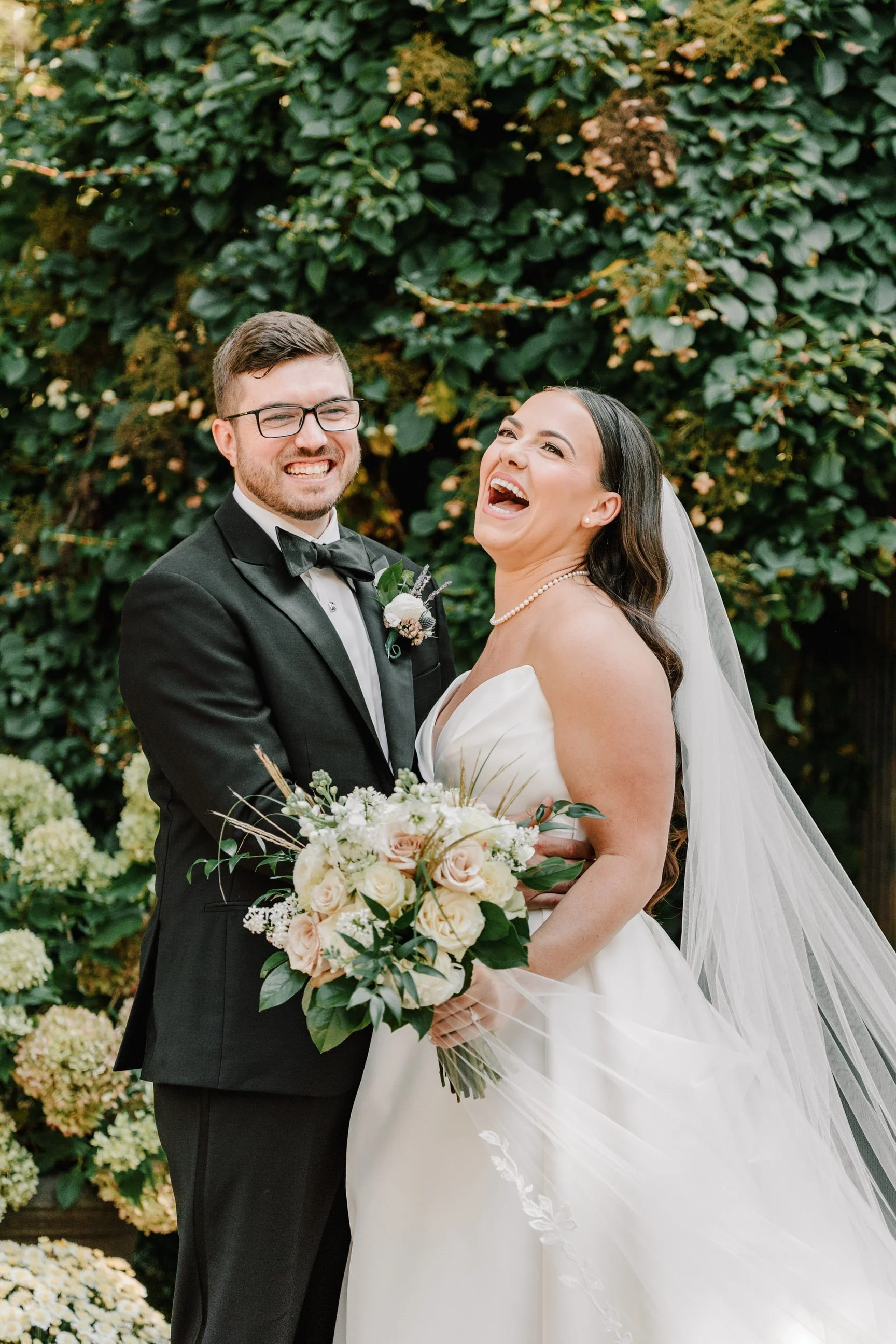 A happy bride and groom stand outdoors, smiling and laughing. The groom wears a black tuxedo with a bow tie and glasses, while the bride wears a white wedding dress with a pearl necklace and veil, holding a bouquet of pink and white roses. They are s
