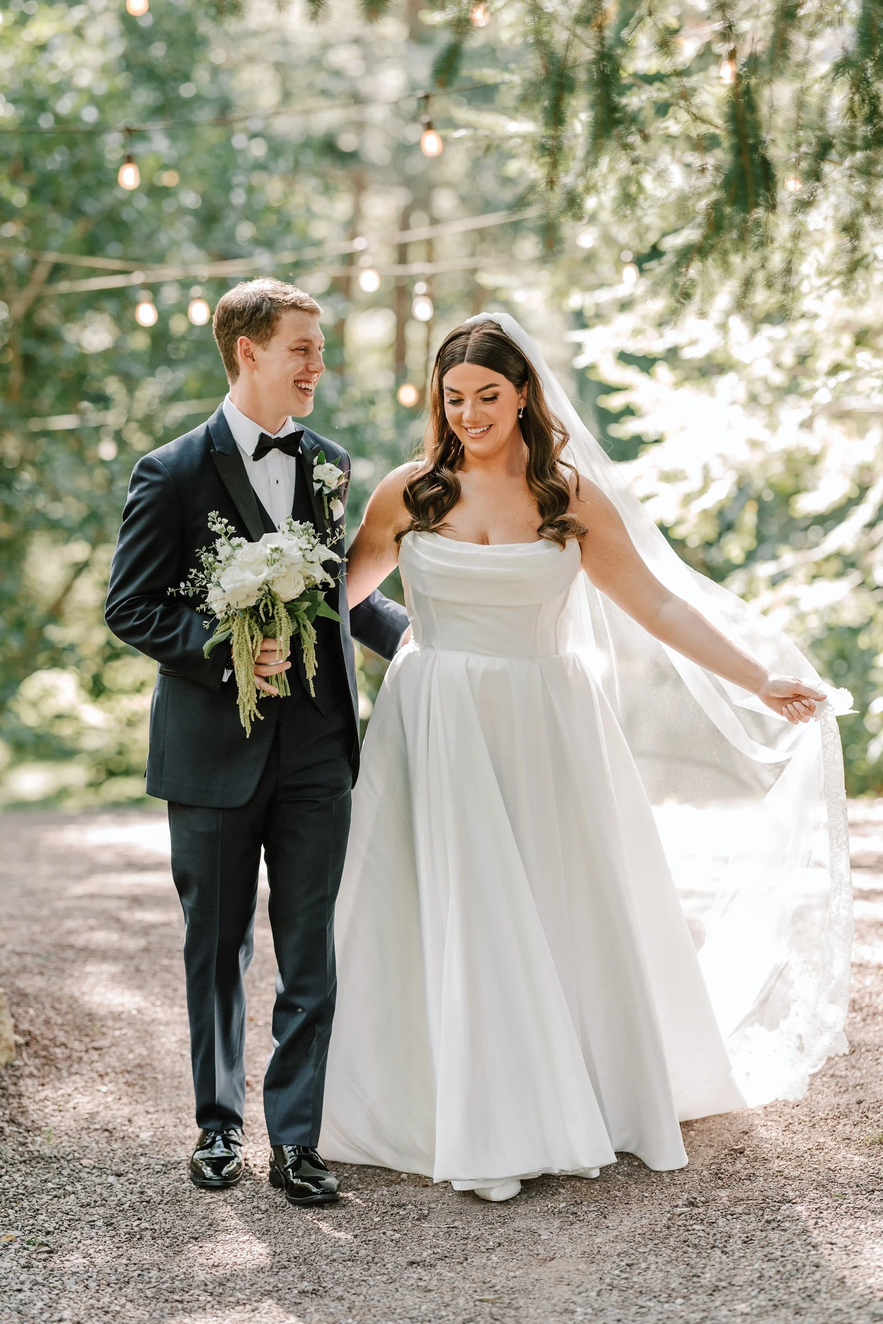 A bride and groom walk outdoors in a wooded area during their wedding, smiling and holding hands. The bride wears a white gown and veil, and the groom wears a black tuxedo with a bow tie, holding a bouquet of white flowers. String lights hang above t