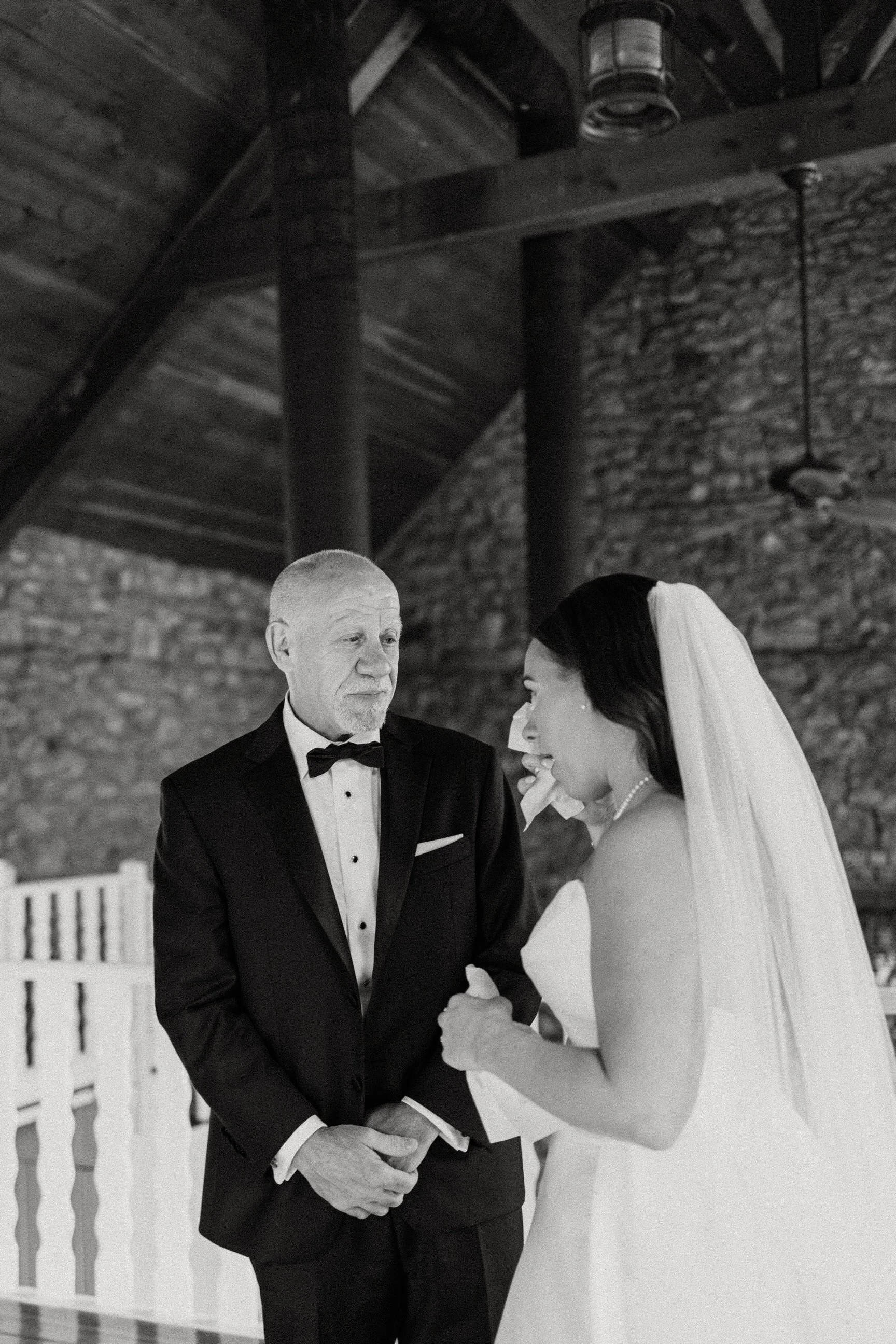 An elderly man in a tuxedo and a woman in a wedding dress holding hands during a wedding ceremony in a rustic venue with exposed brick walls and wooden beams.