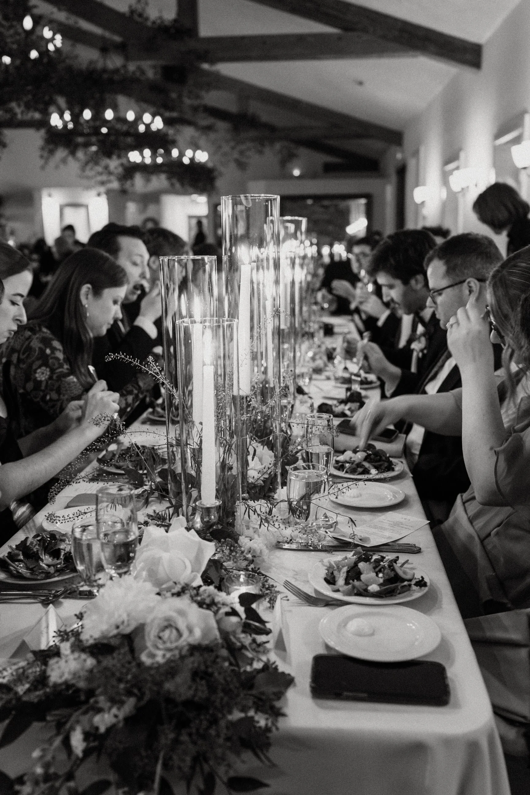 A black and white photo of a formal dinner event with people seated at a long table, decorated with tall glass candle holders and floral arrangements.