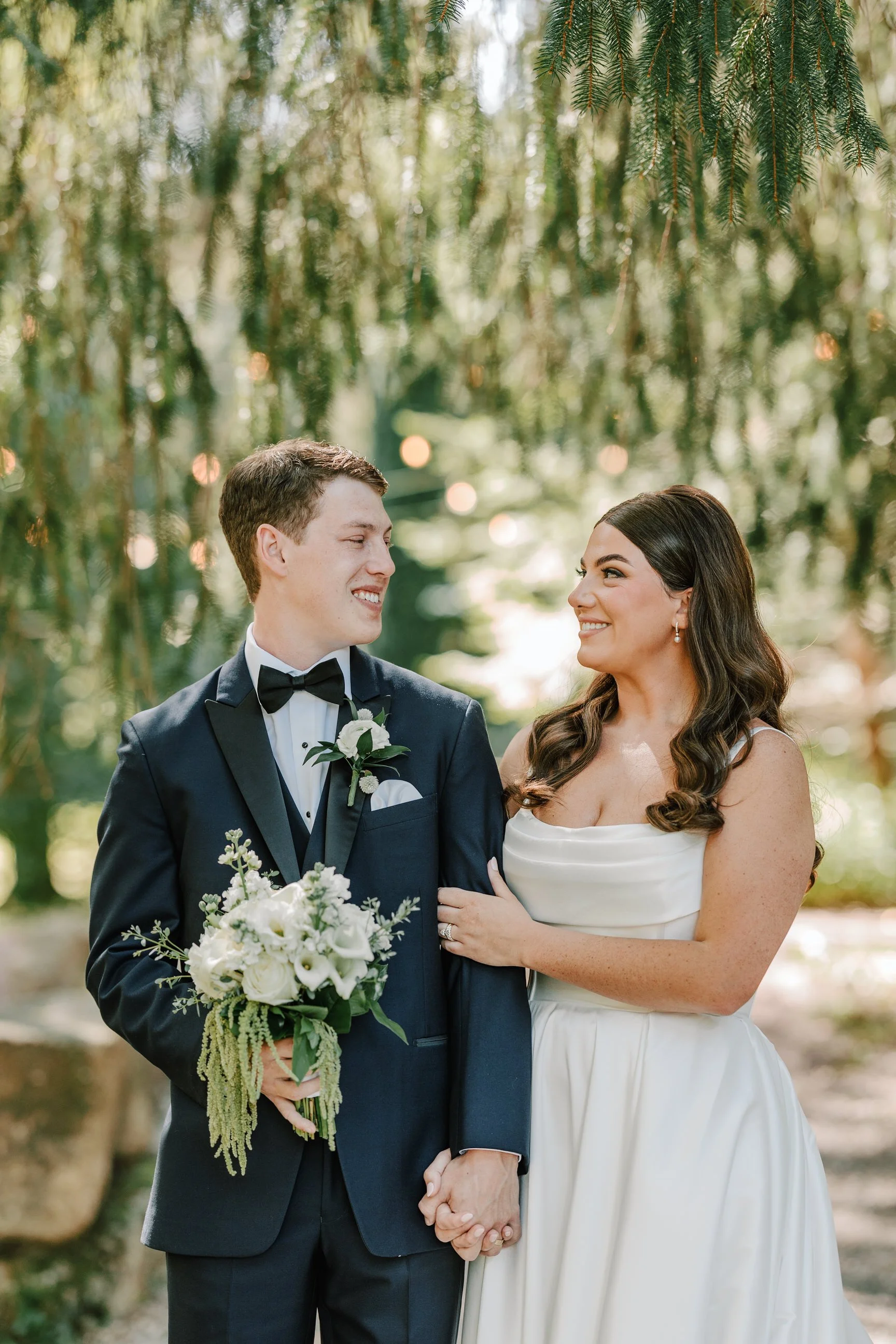A newlywed couple holding hands and smiling at each other outdoors. The groom is dressed in a dark tuxedo with a black bow tie and a white boutonniere. The bride is wearing a strapless white wedding gown and pearl earrings, holding a bouquet of white