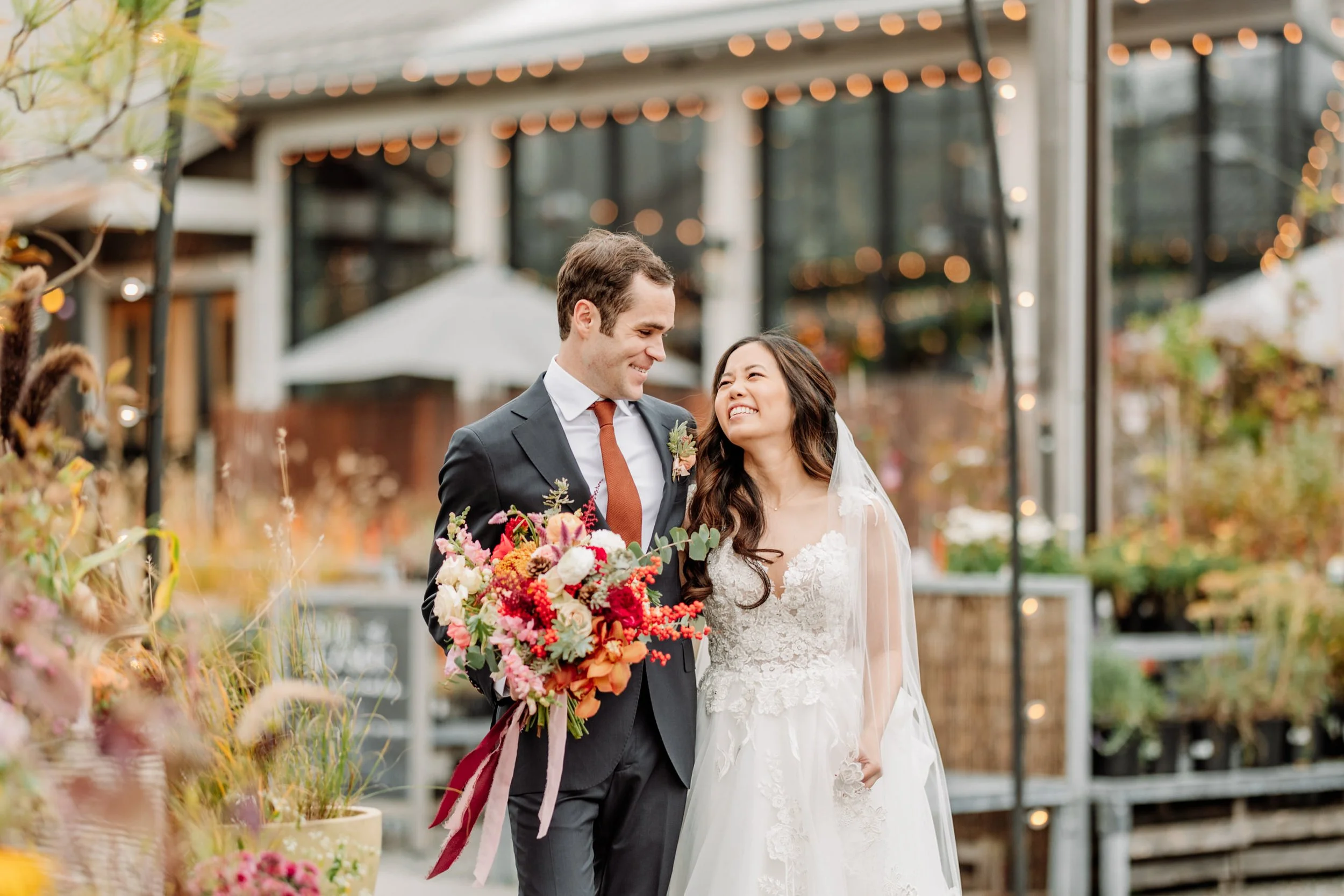 Bride and groom at wedding, smiling at each other, outdoor setting with string lights, flowers, and patio background.
