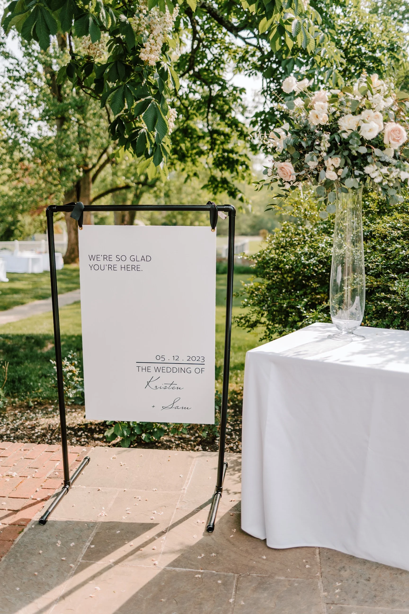 Wedding welcome sign on a stand with a white background and black text, next to a table with a white cloth and a tall glass vase with pink and white flowers outdoors.