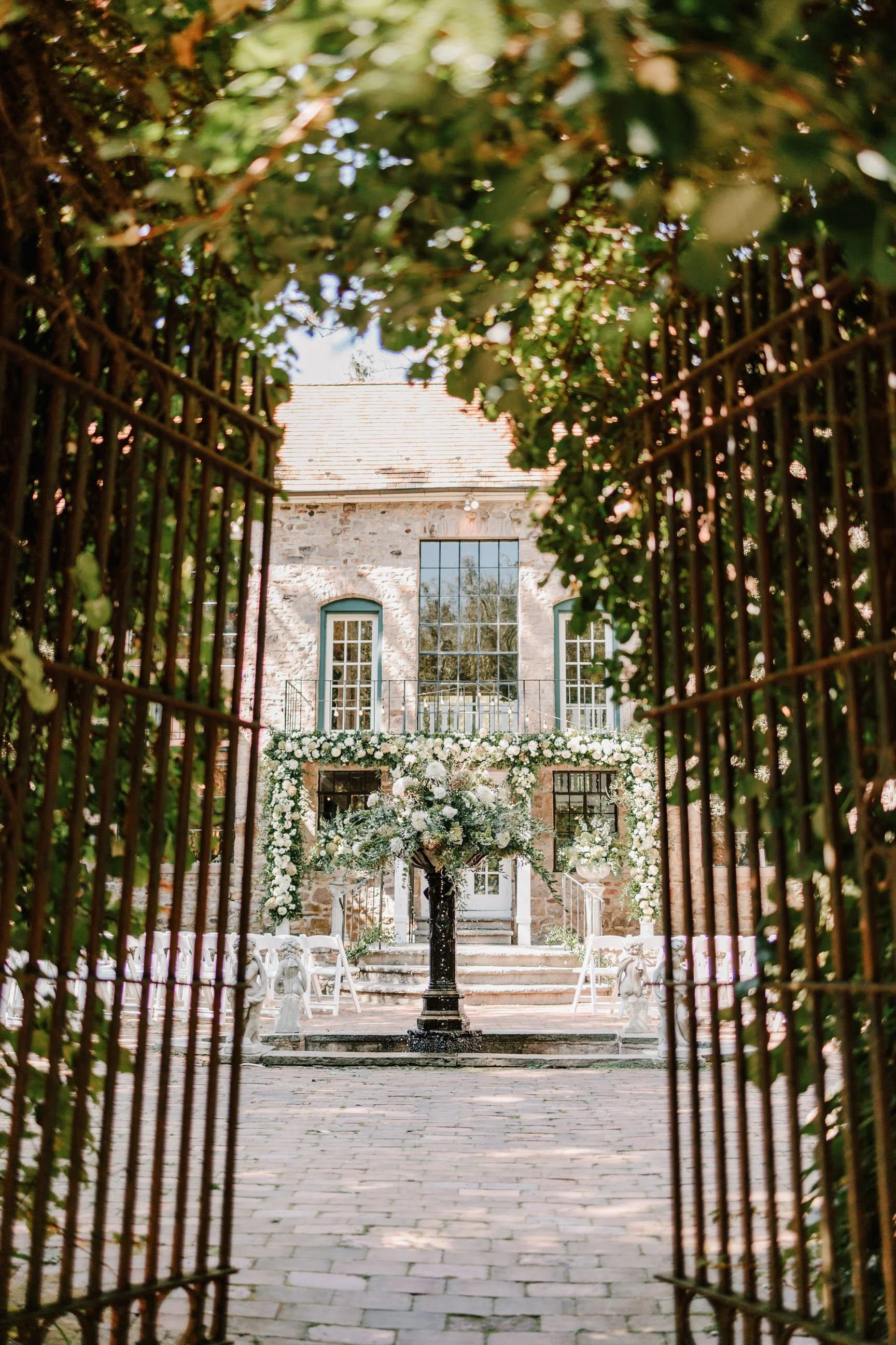 A wedding venue with a stone building decorated with white flowers, viewed through a metal gate and greenery.