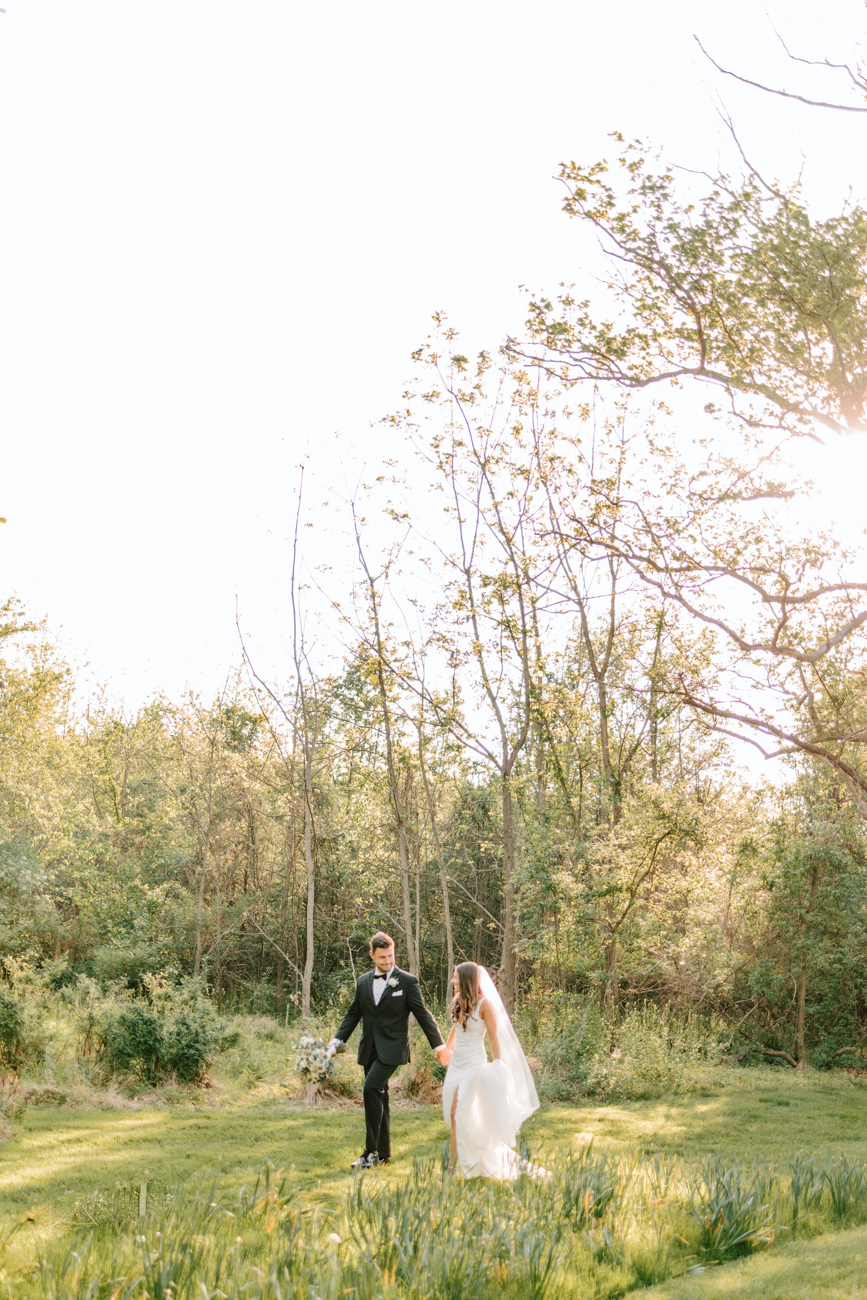 A newlywed couple holding hands and walking through a lush green garden with tall trees, under a bright sky with the sun shining.