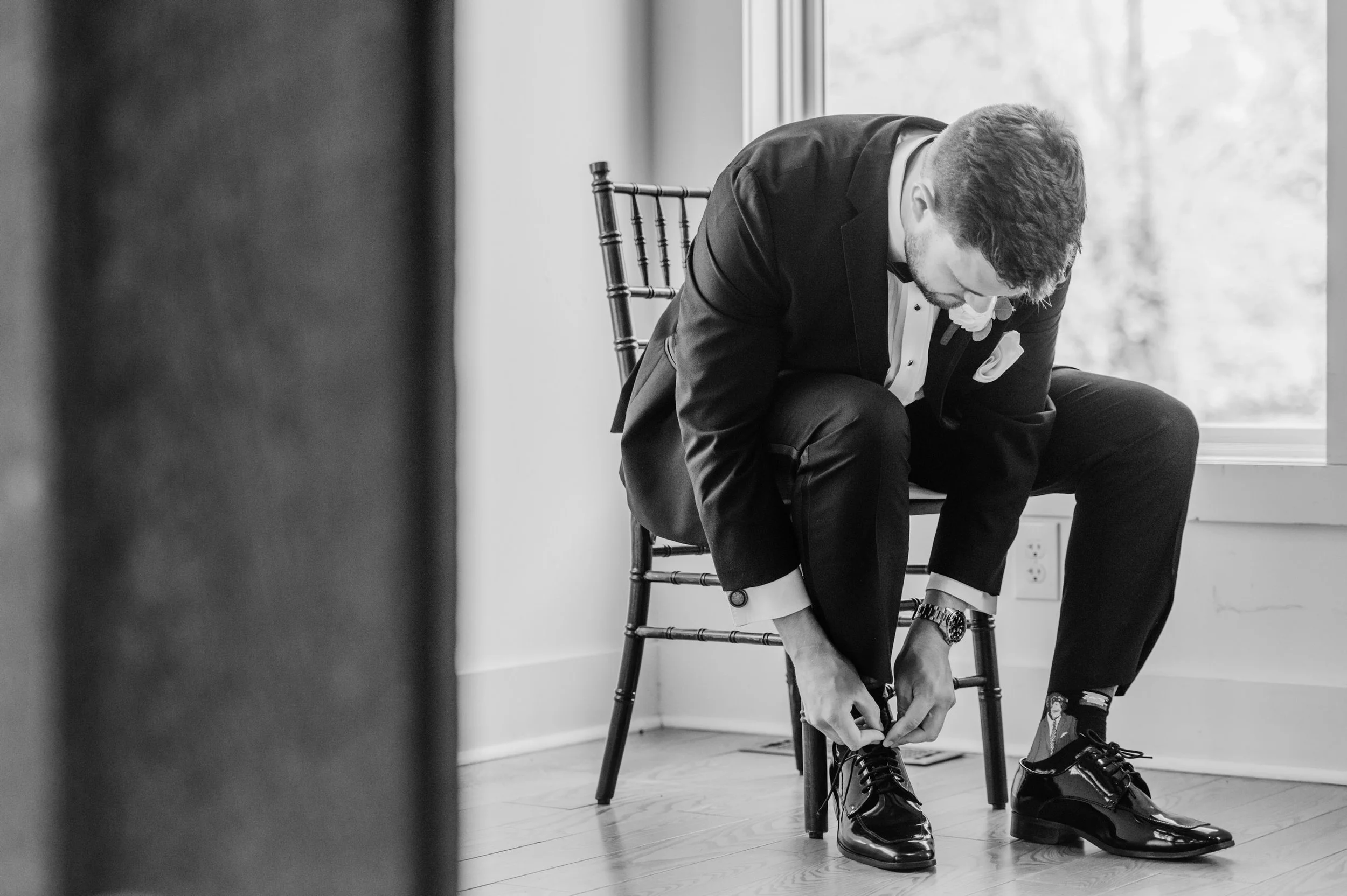 A man in a tuxedo sits on a chair, tying his shoelaces, with a window in the background showing trees outside.