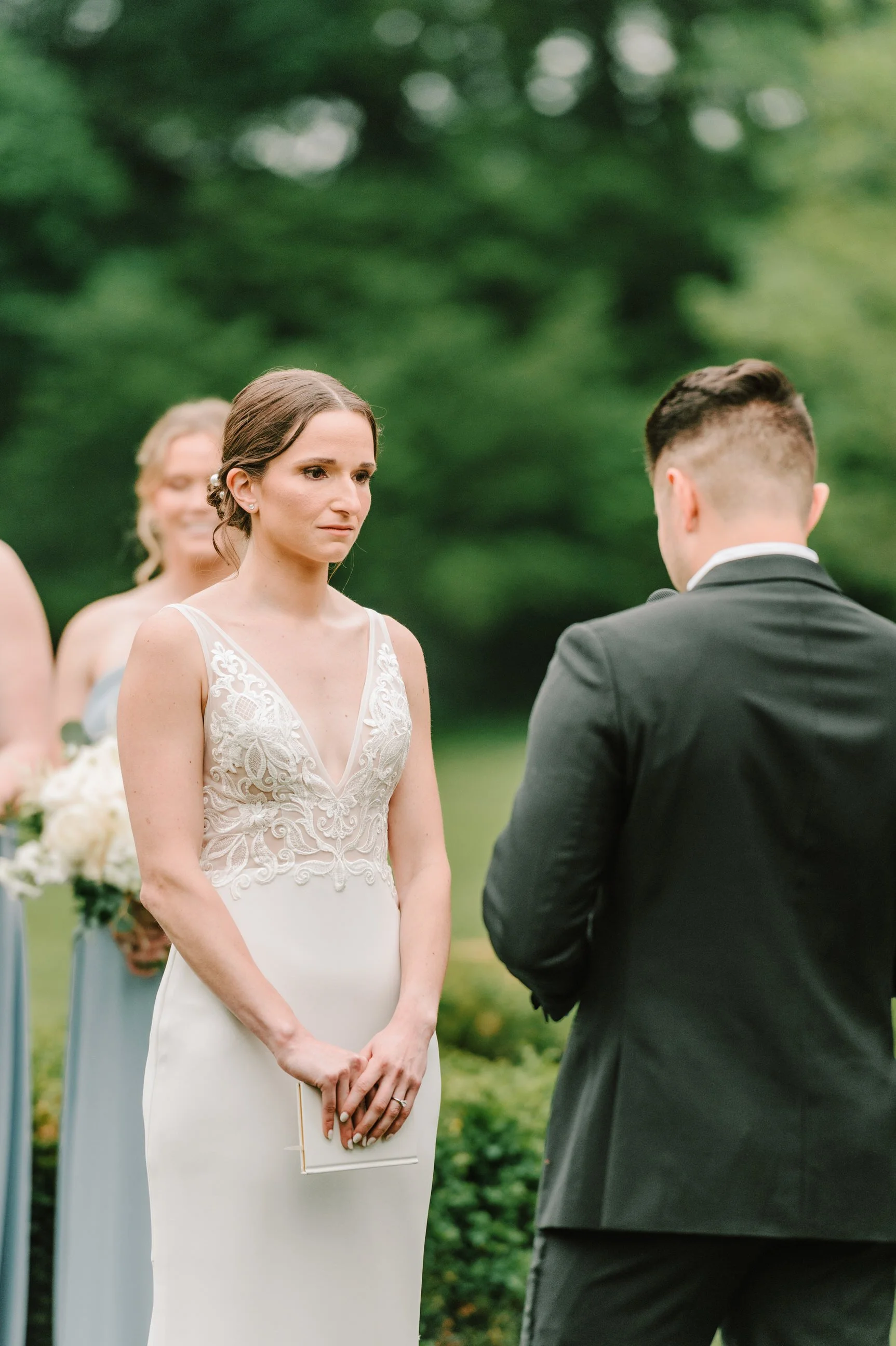 A woman in a wedding dress and a man in a suit stand outdoors during a wedding ceremony, facing each other.