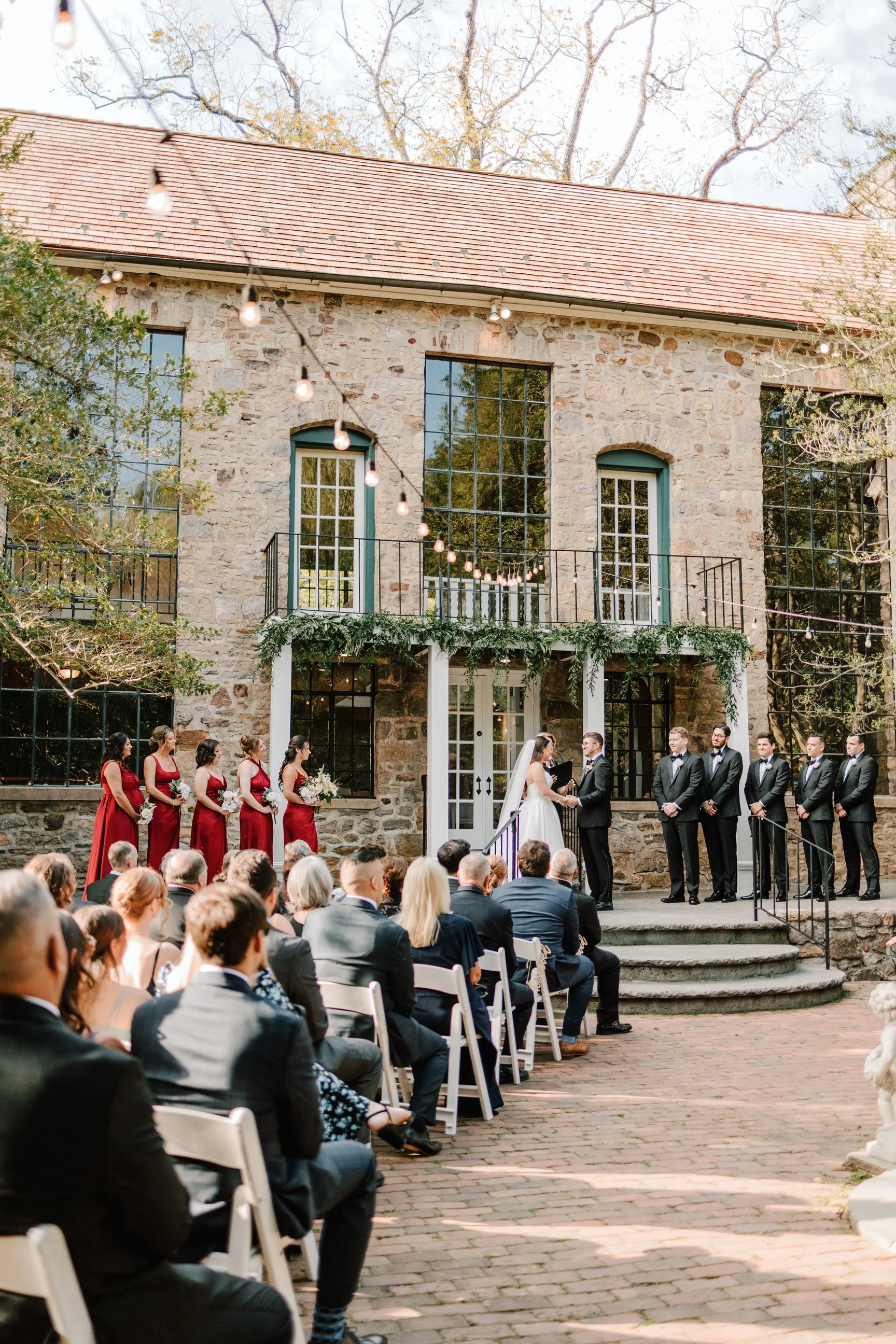 A wedding ceremony taking place outdoors with the bride and groom exchanging vows in front of a stone building, surrounded by guests seated in white chairs, with bridesmaids in red dresses and groomsmen in black tuxedos. String lights are hanging ove