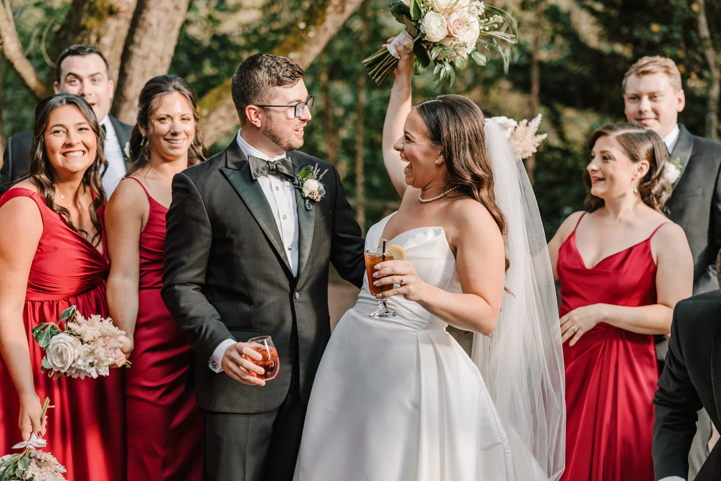 A wedding celebration with the bride in a white gown and the groom in a black tuxedo surrounded by bridesmaids in red dresses and groomsmen in black suits, outdoors with trees in the background.
