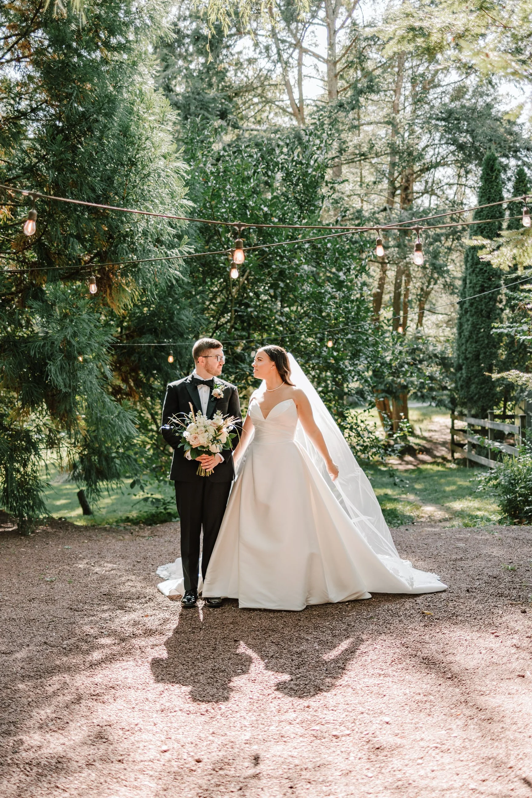 Bride and groom standing outdoors, holding a bouquet of flowers, under string lights amidst trees in a scenic setting.