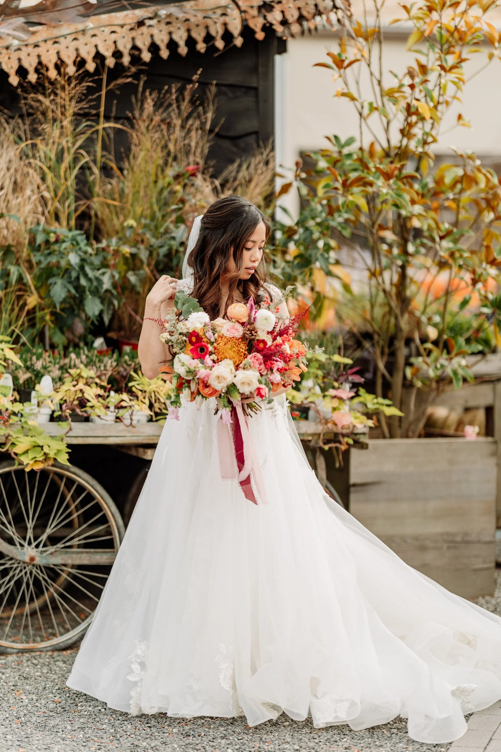 A bride in a white wedding dress holding a large bouquet of colorful flowers, standing outdoors near a wooden cart with plants and autumn foliage in the background.