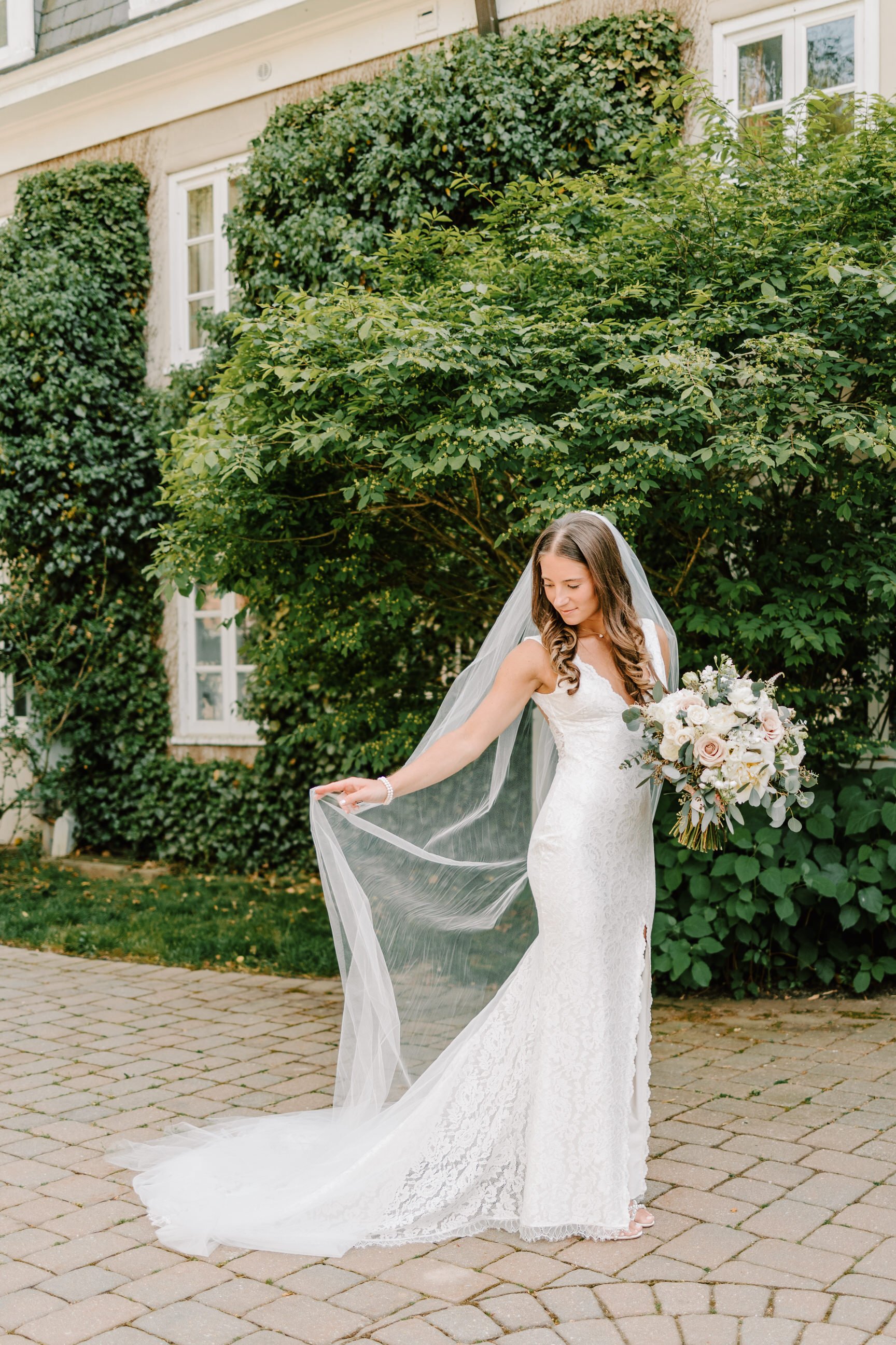 Bride in a white lace wedding dress holding a bouquet while standing on a brick pathway outdoors, with green bushes and a house in the background.