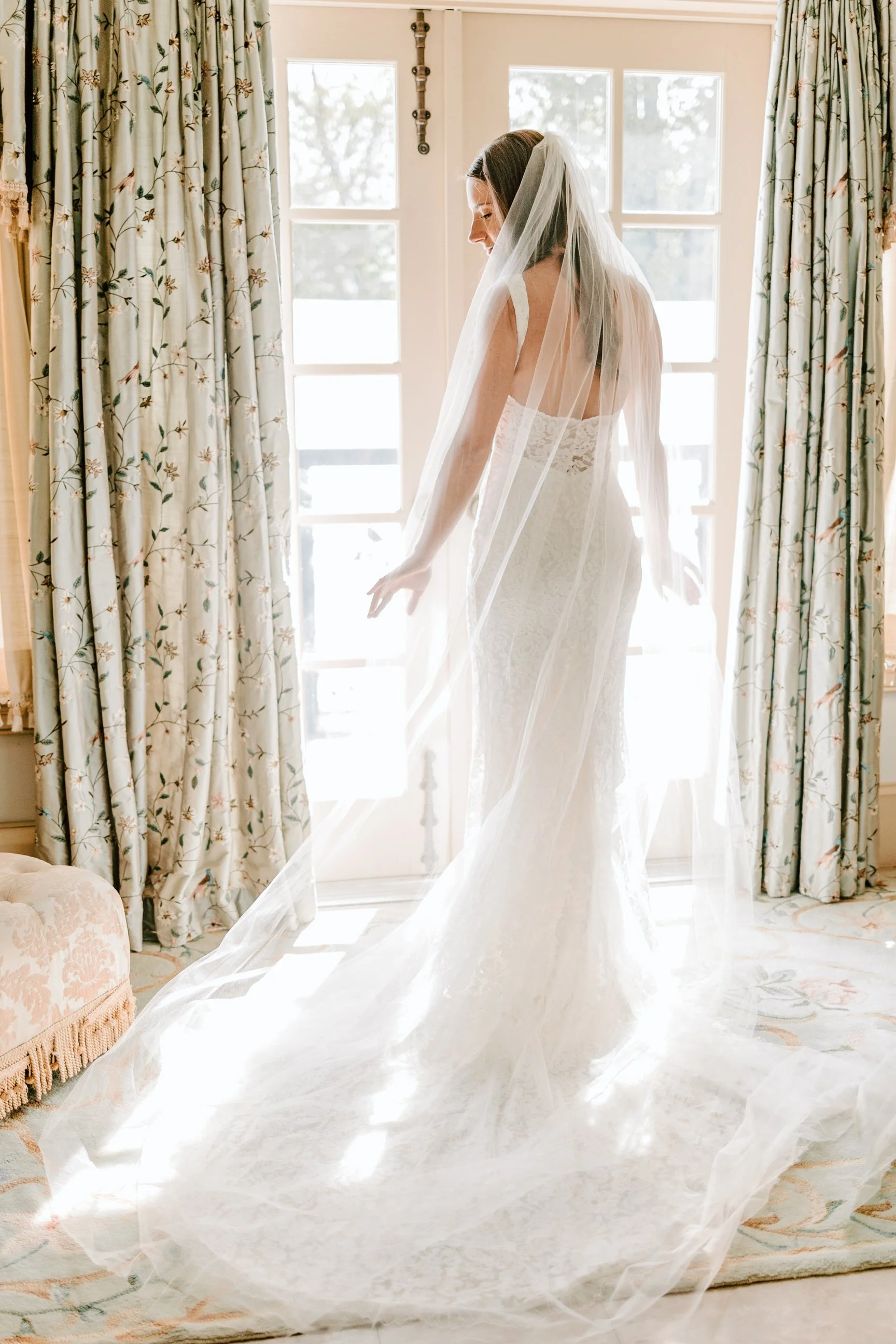 Bride in a lace wedding dress and veil standing by a window, with sunlight illuminating her and floral curtains in the background.