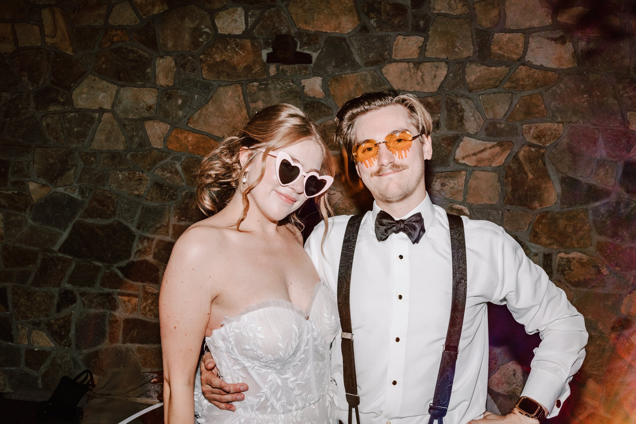 A smiling couple at a party, the woman wearing heart-shaped sunglasses and a strapless white dress, the man wearing orange-tinted glasses, a white shirt, black suspenders, a bow tie, and a smartwatch, standing in front of a stone wall.