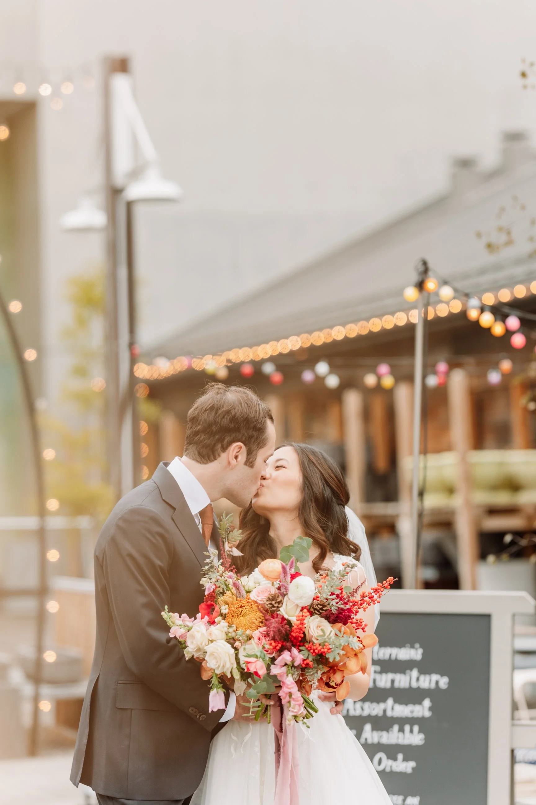 A bride and groom share a kiss during their wedding, with the bride holding a bouquet of colorful flowers, in an outdoor setting decorated with string lights.