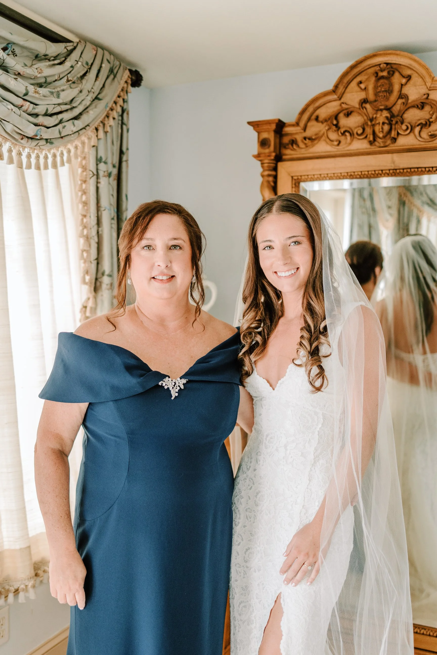 A woman in a navy blue dress standing next to a bride in a white lace wedding gown with a veil. They are indoors near a mirror with a wooden frame, in a room with decorated curtains and light-colored walls.