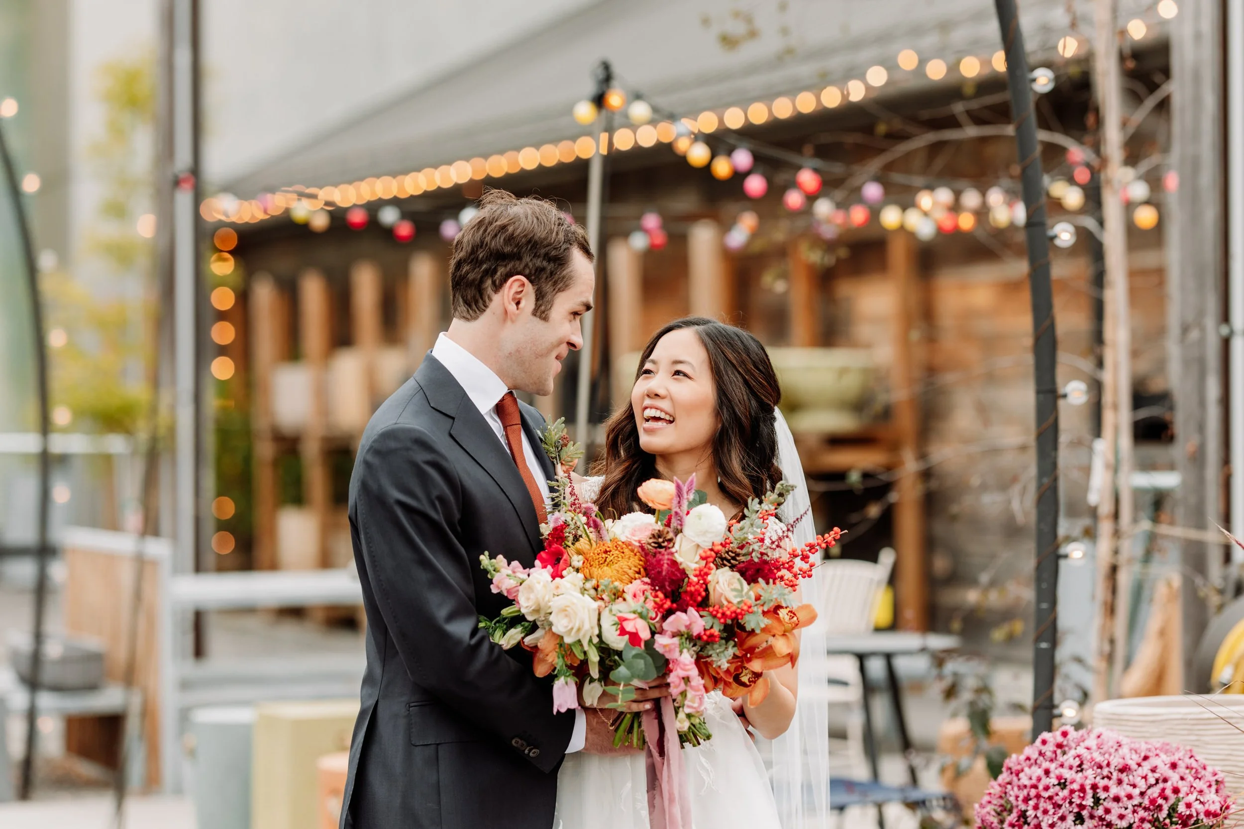 A bride and groom holding a bouquet of flowers and looking at each other at their wedding reception outdoors, with string lights and wooden decor in the background.