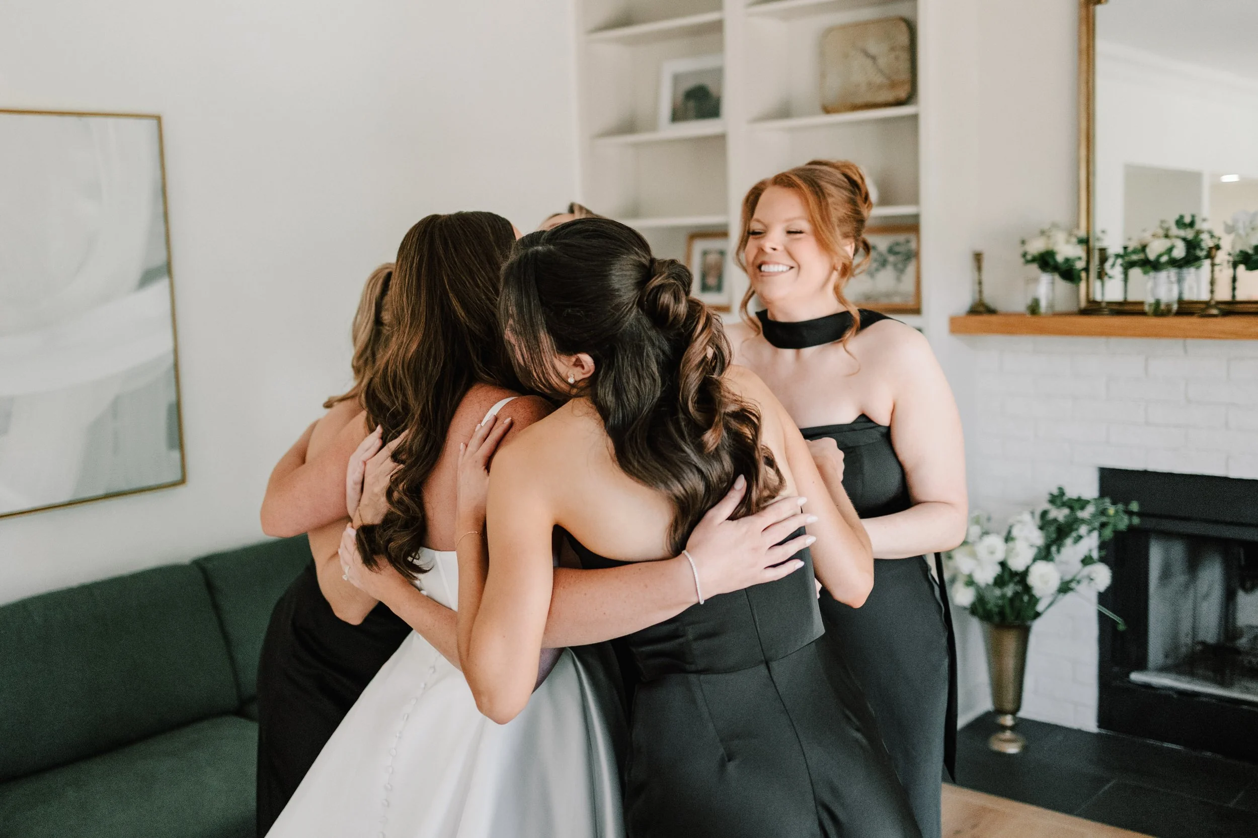 Group of women embracing and smiling in a cozy living room, with a white brick fireplace, framed photos, and plants in the background.