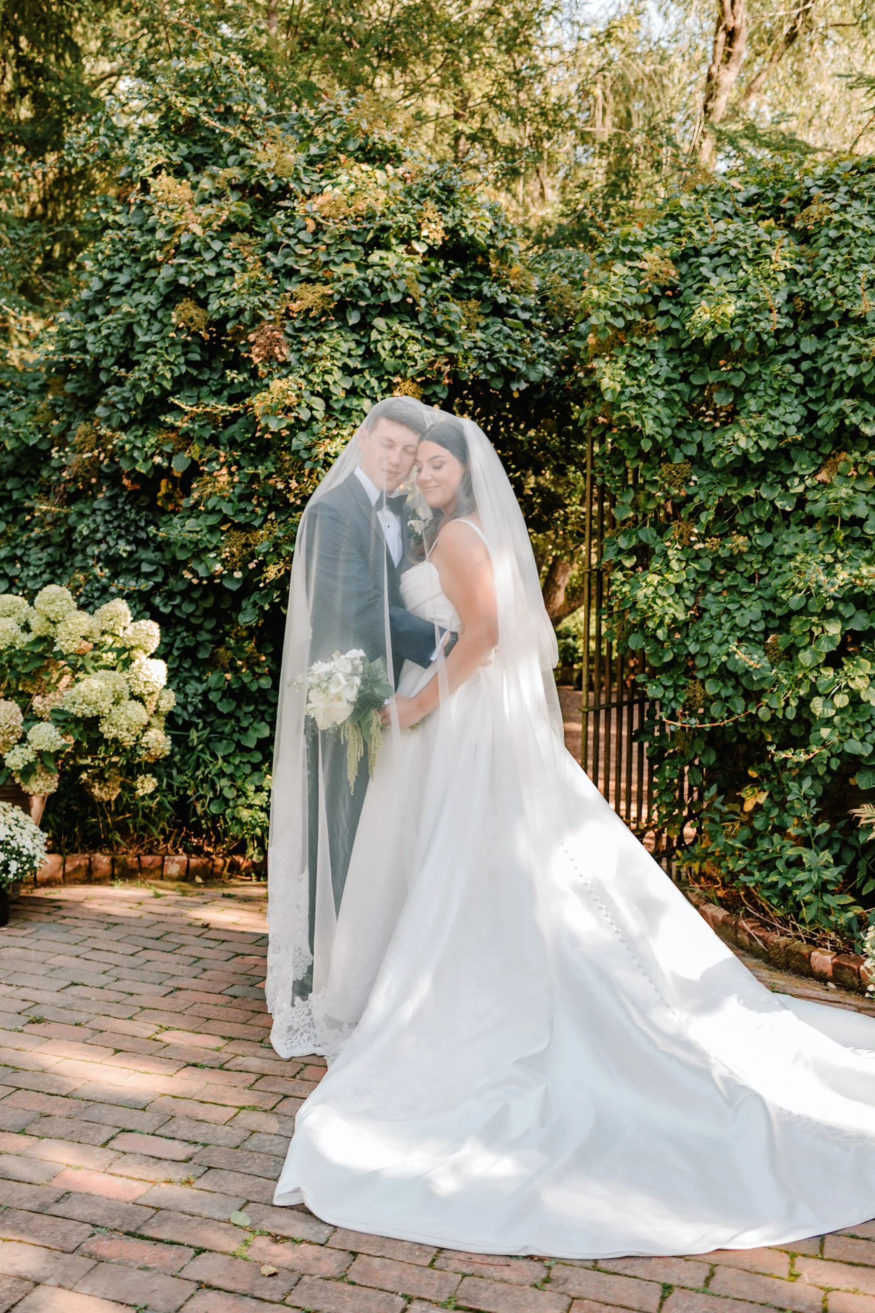 A bride and groom standing under a wedding veil outdoors, with bushes and trees in the background, on a brick pathway.