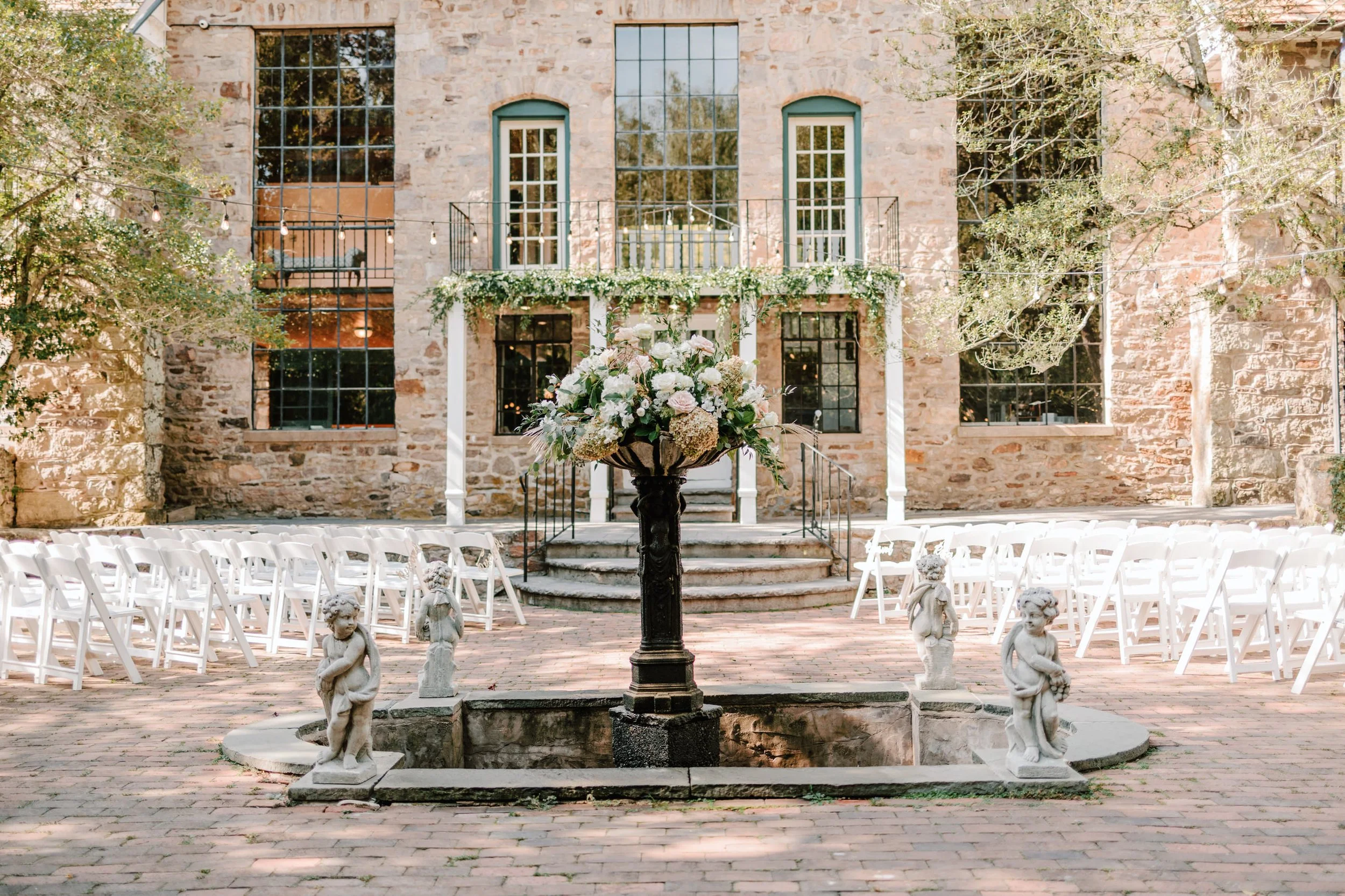 Outdoor wedding ceremony setup with white chairs, a flower arrangement on a central fountain, and a stone building in the background.