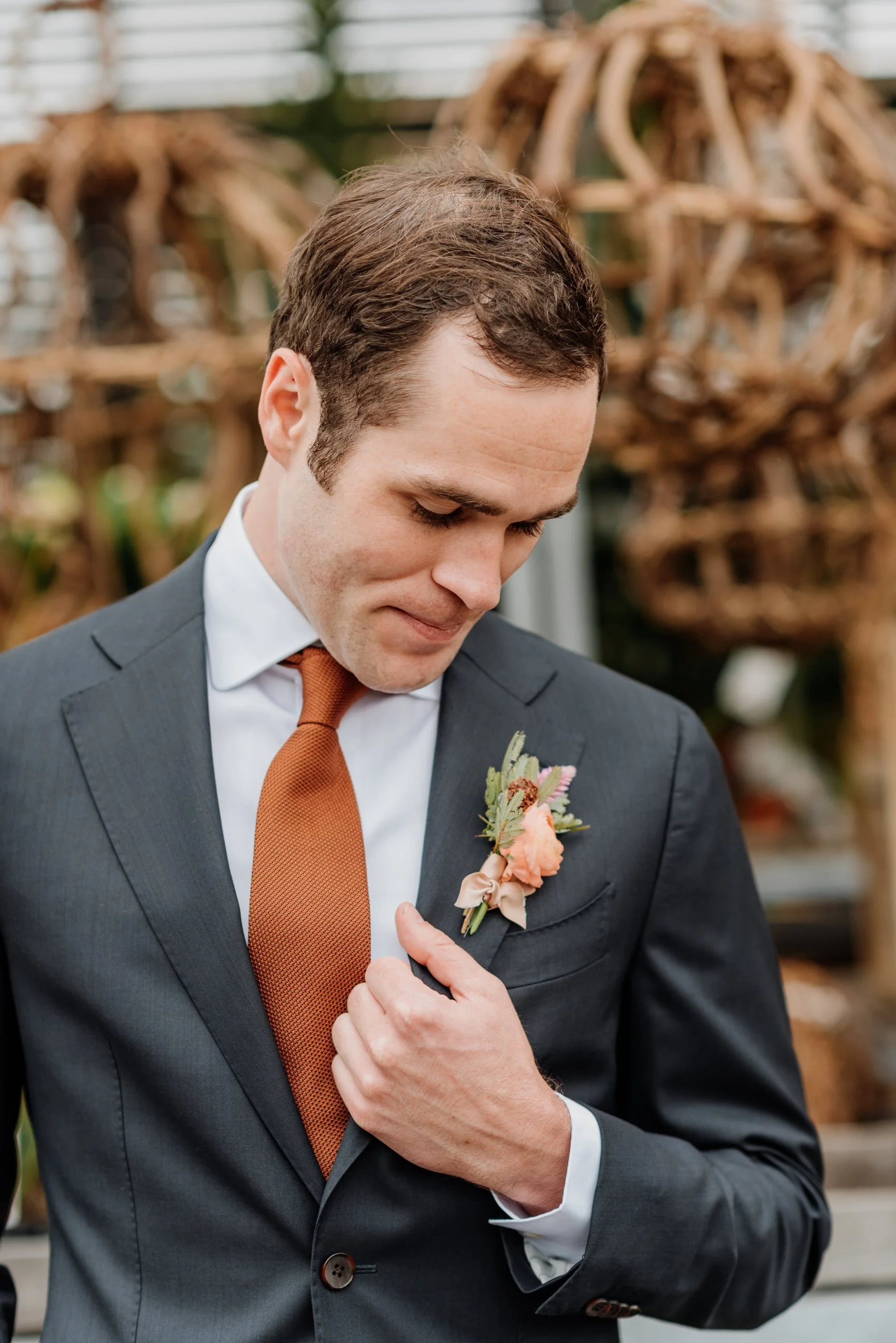A man in a dark gray suit with a white shirt and a rust-colored tie, holding his lapel and looking down, with a pink floral boutonniere on his lapel, indoors with a rustic decorative background.