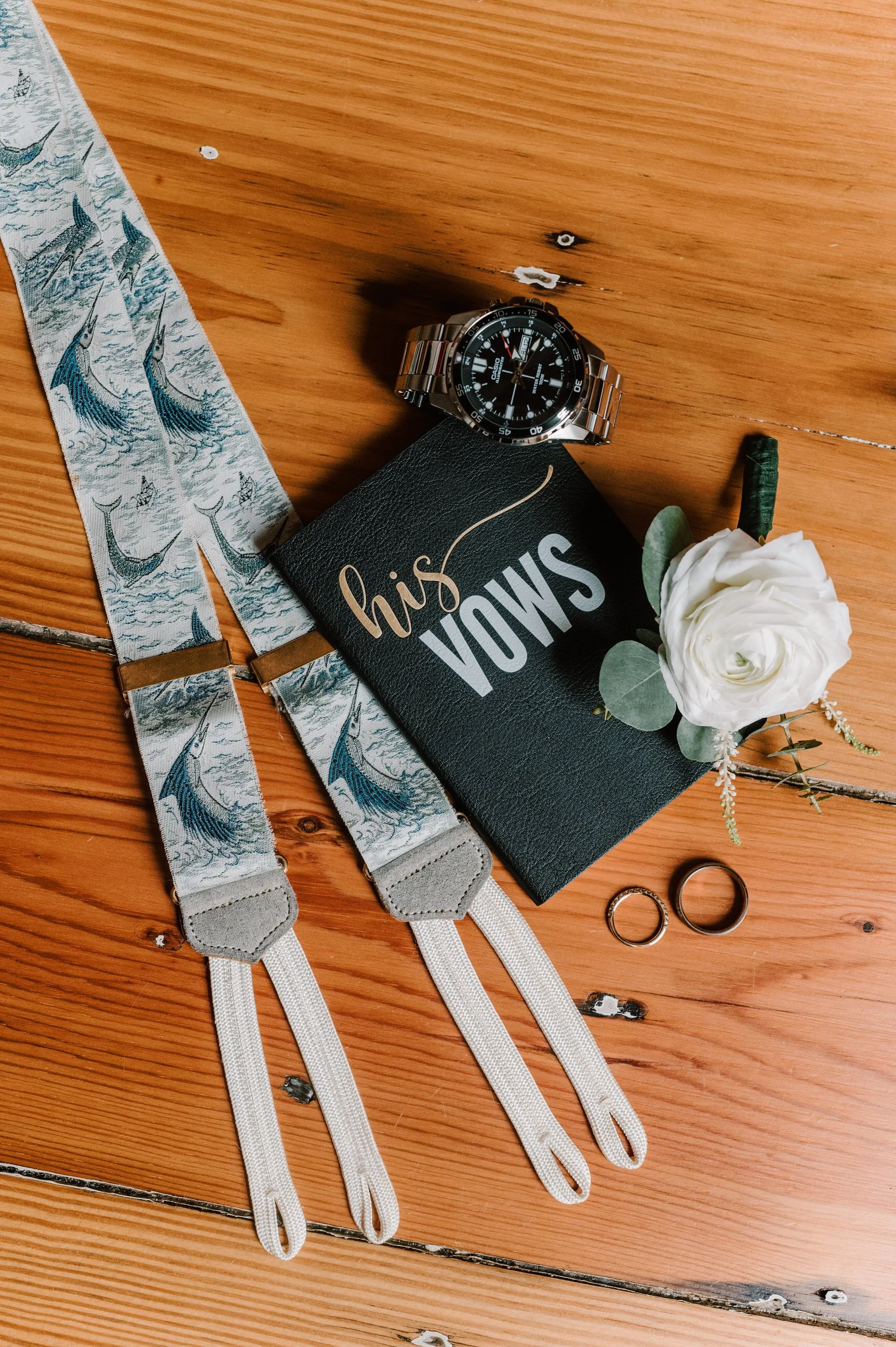 Wedding rings, a floral boutonniere, a wristwatch, a black notebook with 'his VOWS' written on it, and suspenders with a whale pattern on a wooden surface.
