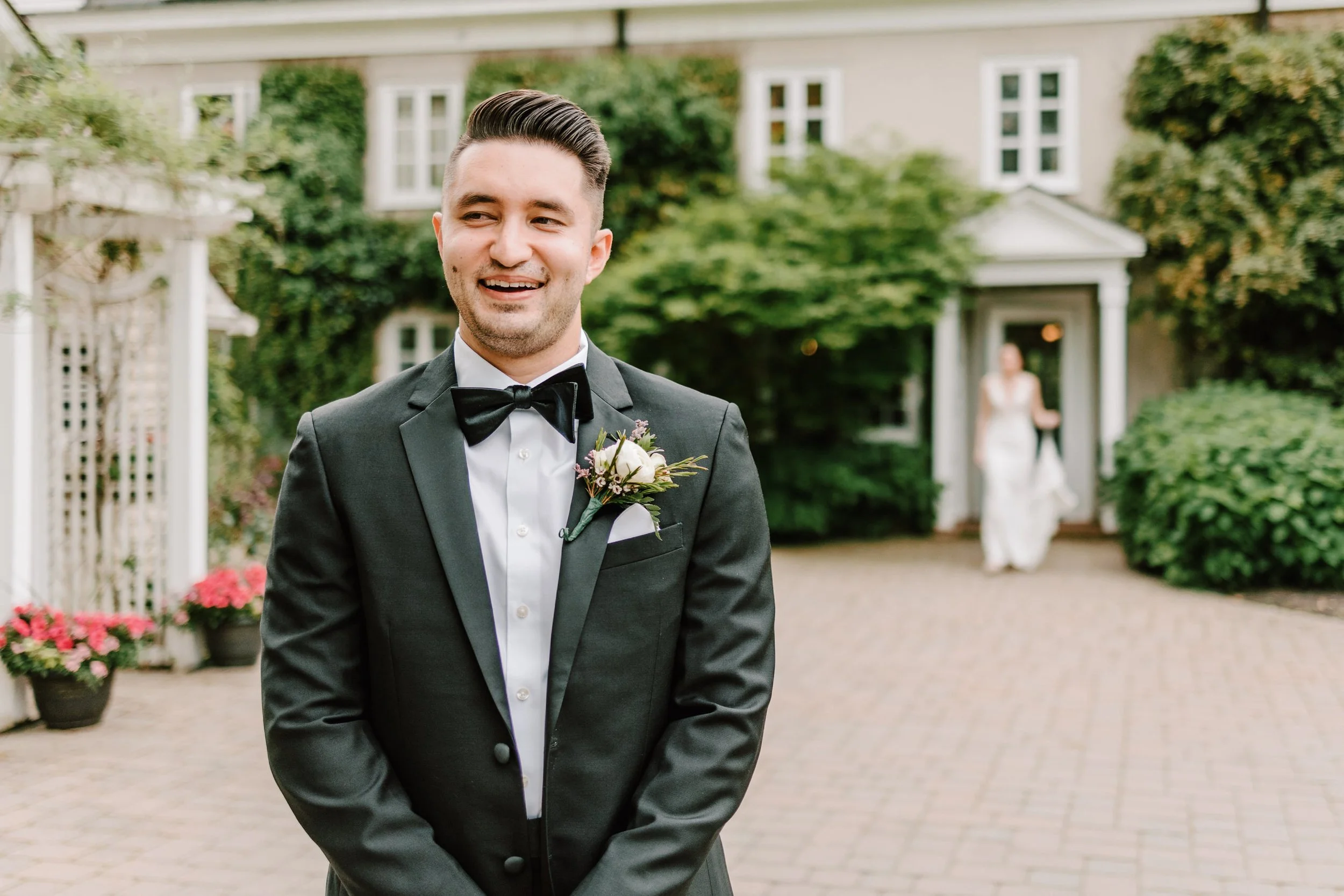 A groom in a black tuxedo with a bow tie and boutonniere, smiling outdoors in front of a house with greenery and pink flowers, with a bride in a white wedding dress walking towards him in the background.