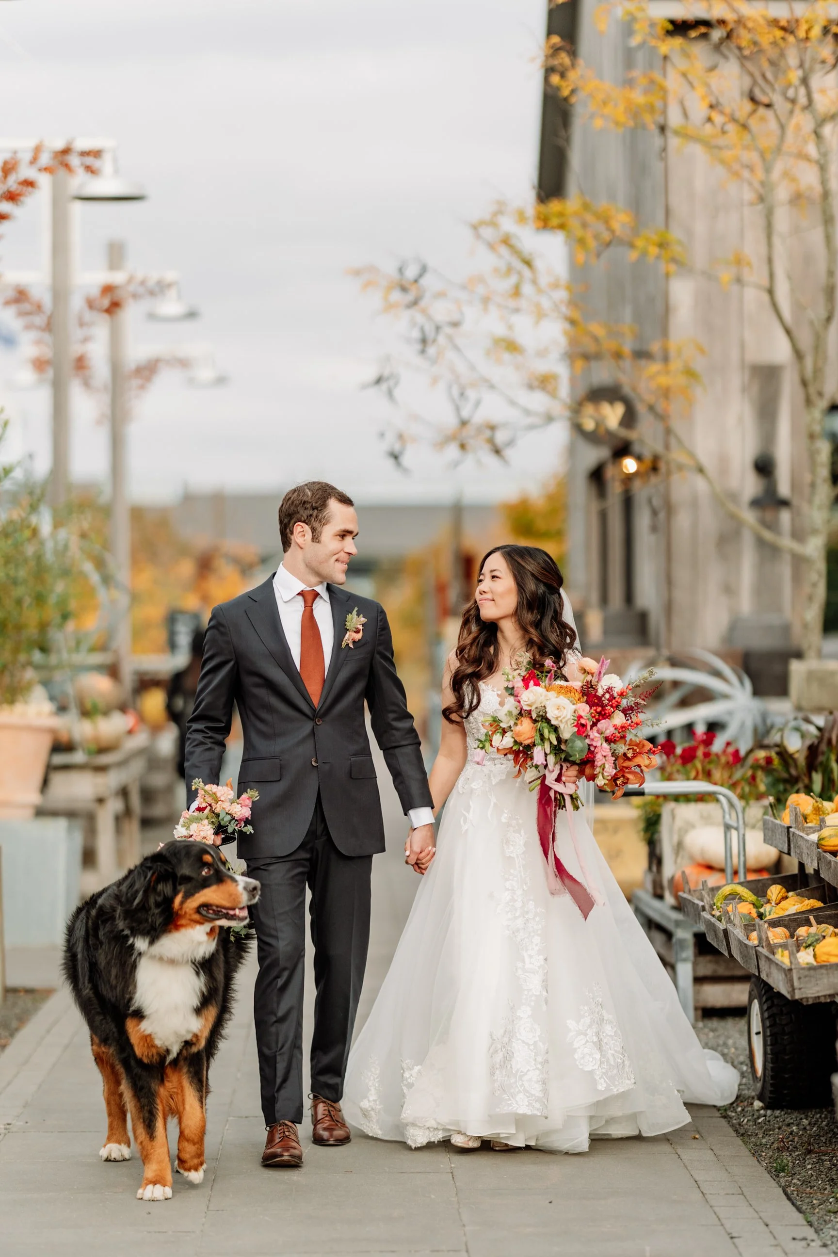 Newlywed couple walking hand-in-hand outdoors with their dog. The bride is holding a bouquet of colorful flowers, and the groom is dressed in a dark suit with a boutonniere. Autumn scenery surrounds them in a rustic setting.