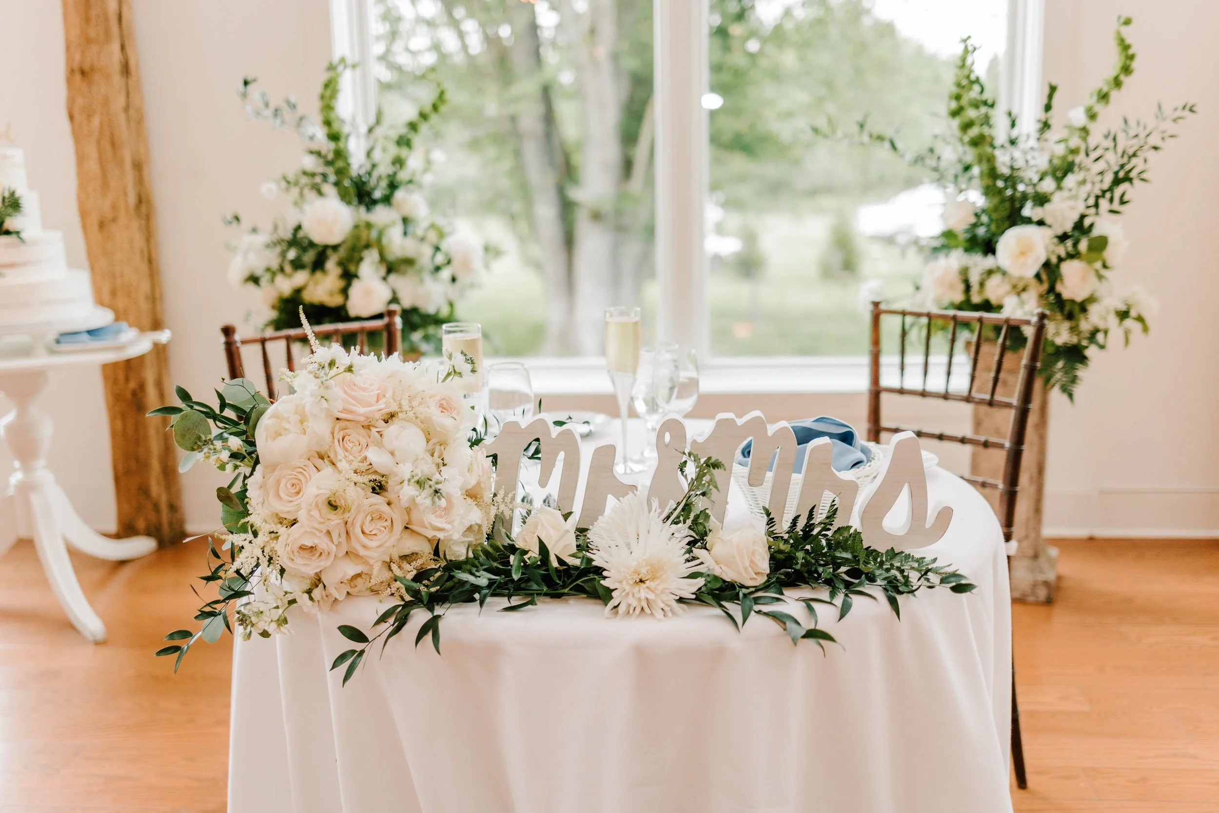 Wedding reception table with floral arrangements, greenery, champagne glasses, and a 'Mr & Mrs' sign. Background features large floral displays and a window with outdoor scenery.