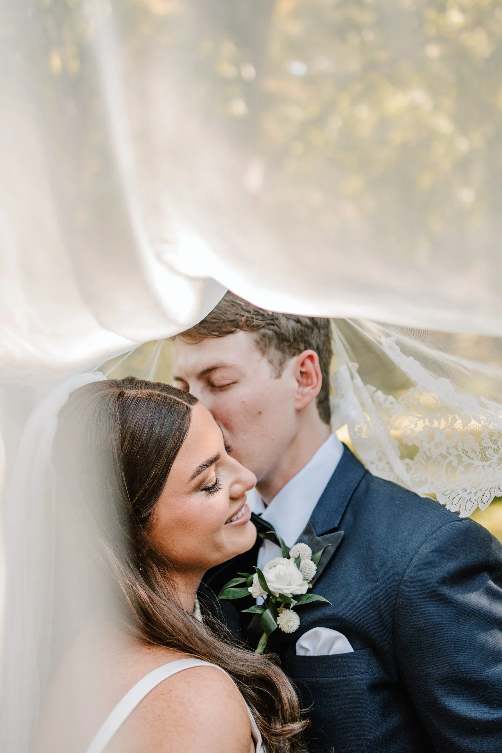 A bride and groom on their wedding day, sharing a tender moment under a veil outdoors.