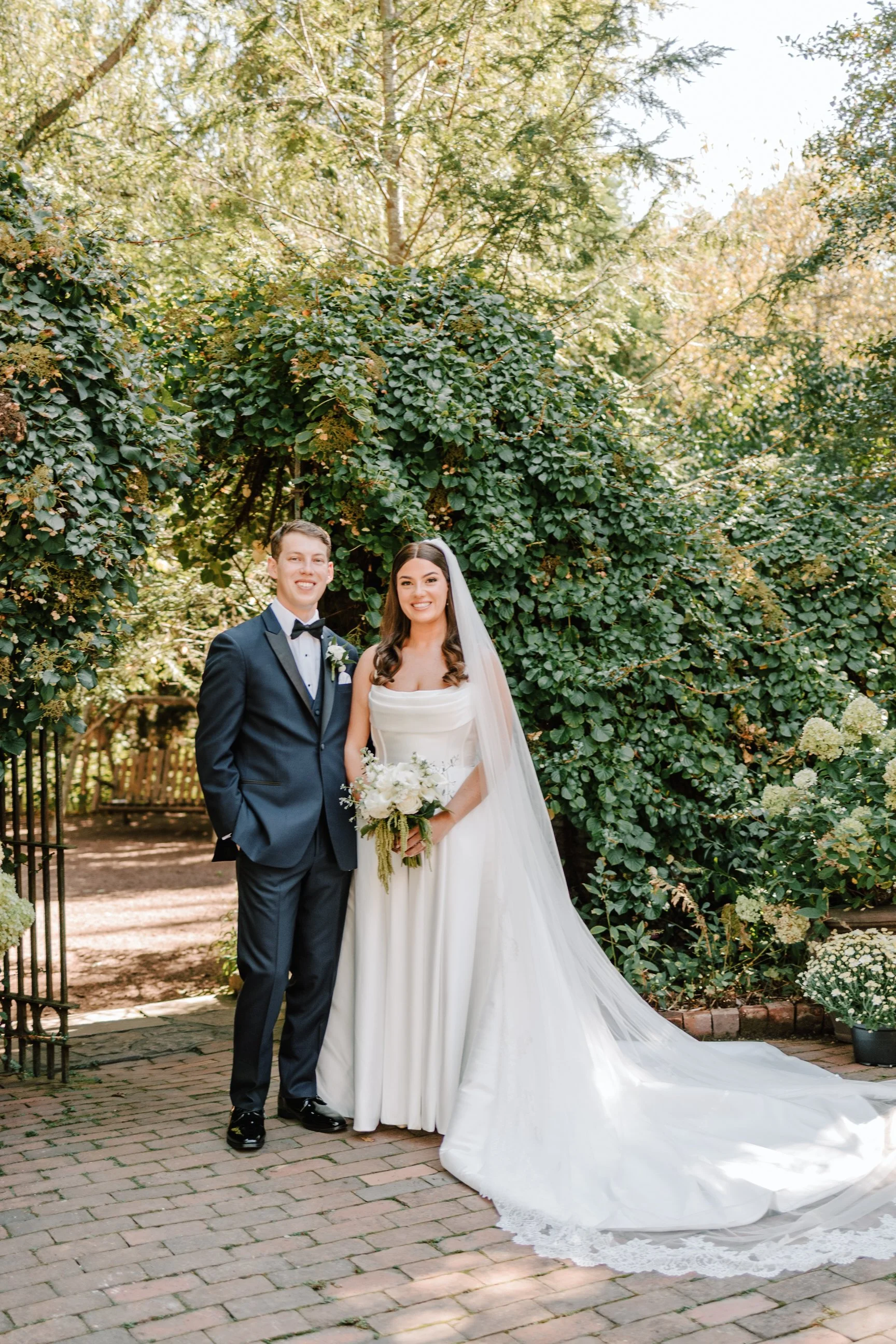 A newlywed couple standing outdoors on a brick pathway, with green trees and bushes behind them. The bride wears a white wedding gown with a long train and veil, holding a bouquet of white flowers. The groom is dressed in a dark suit with a bow tie, 
