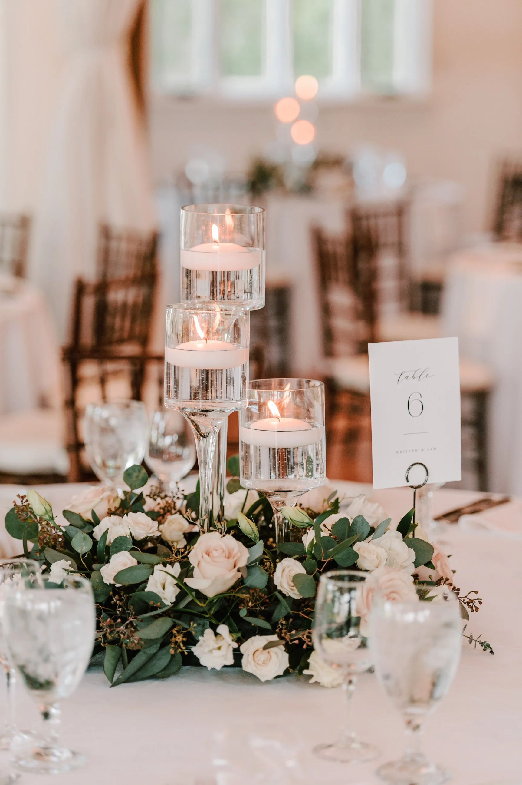 Elegant wedding table centerpiece with three tall floating candles in glass holders, surrounded by white roses and greenery. A table number card indicating 'table 6' is placed among the flowers.