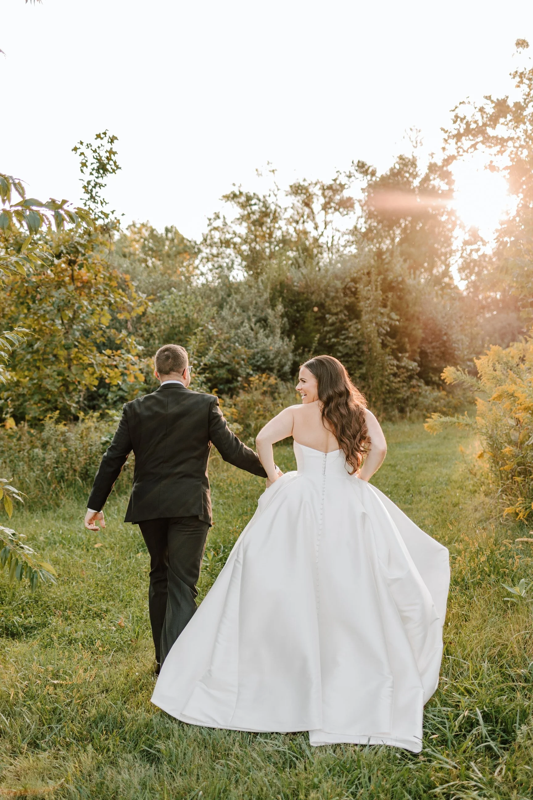 Bride and groom holding hands, walking in a grassy outdoor area at sunset, surrounded by trees and bushes.