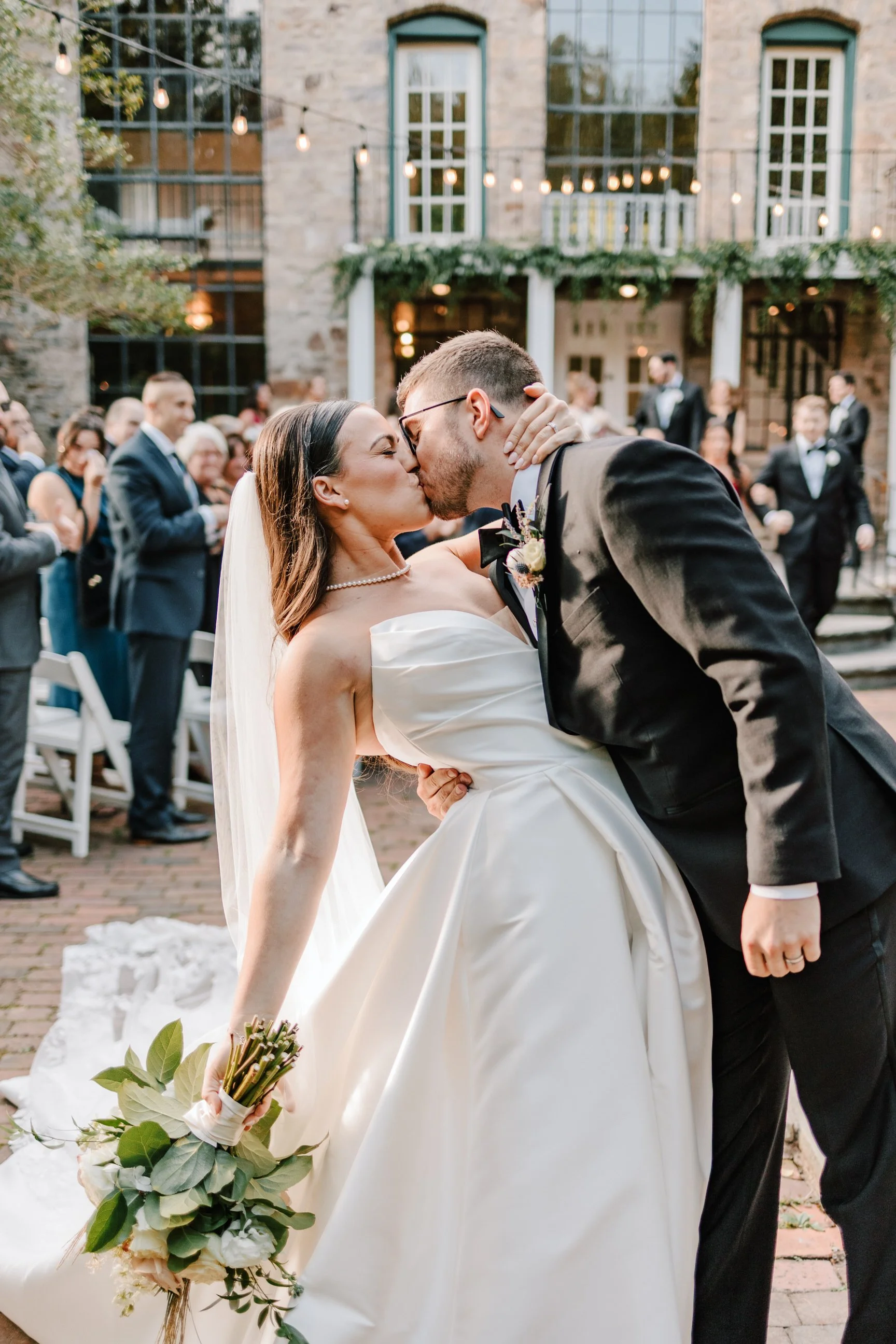 A bride and groom are kissing, with guests in the background during an outdoor wedding ceremony. The bride is wearing a strapless white wedding gown and holding a bouquet, while the groom is in a black tuxedo. There are string lights and a building w