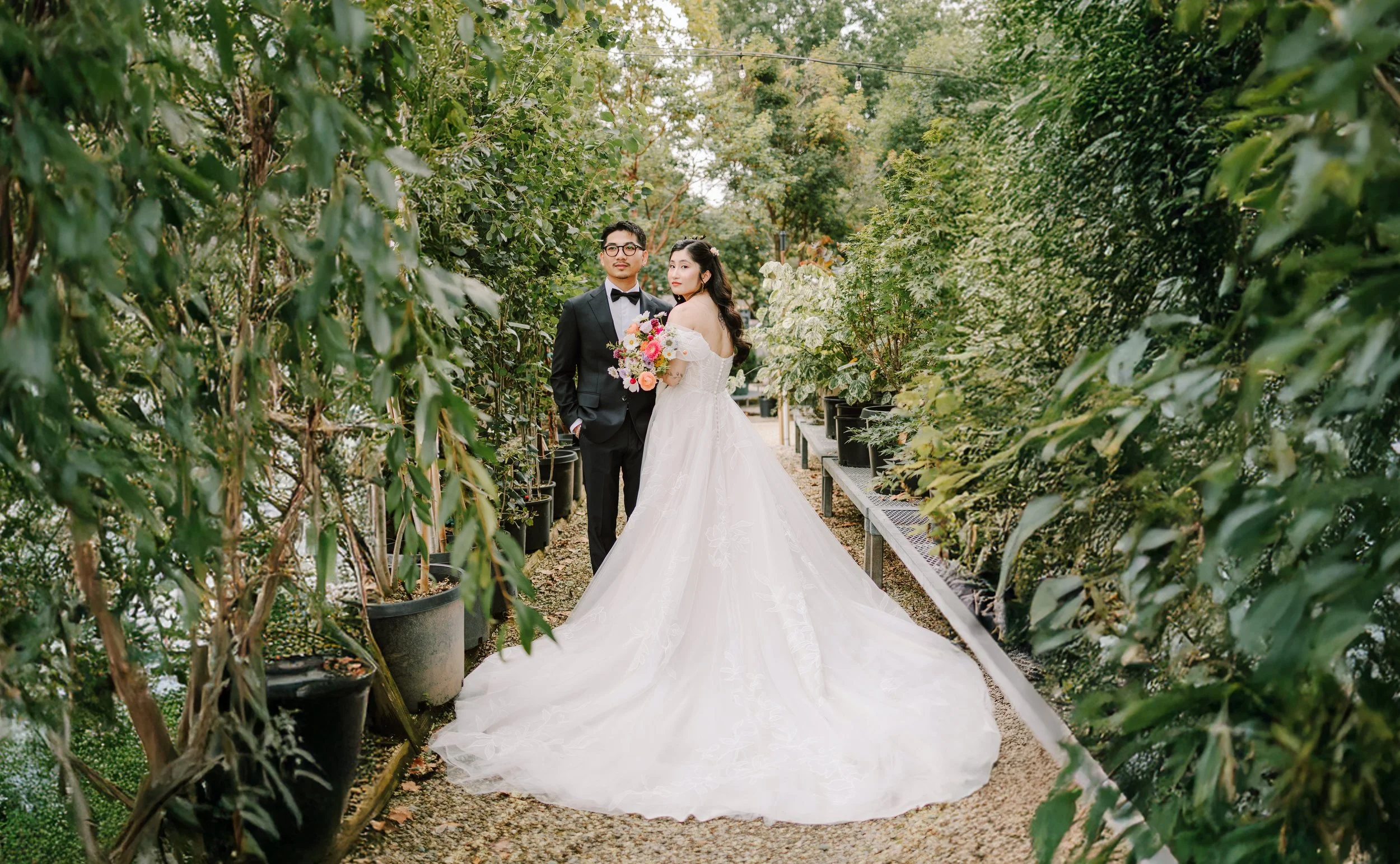 A bride and groom standing together in a lush green garden, the bride holding a bouquet of colorful flowers, both dressed in wedding attire. Philadelphia wedding photographer