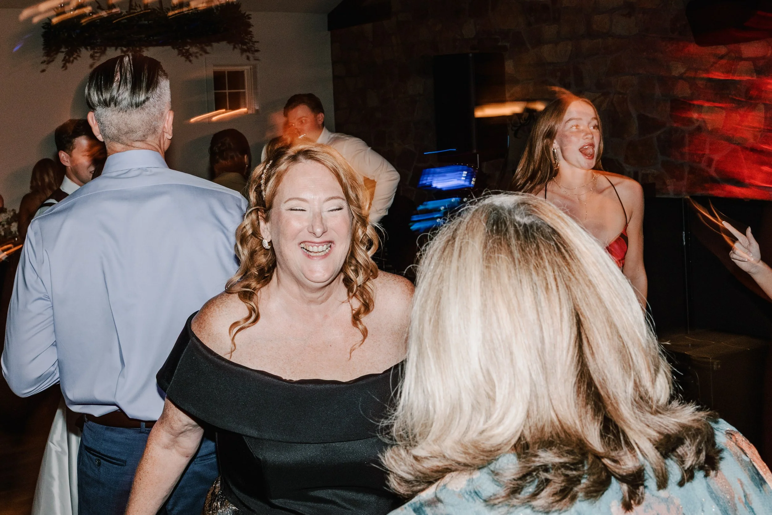 People dancing and laughing at a lively celebration or party, indoors with dim lighting and brick walls.