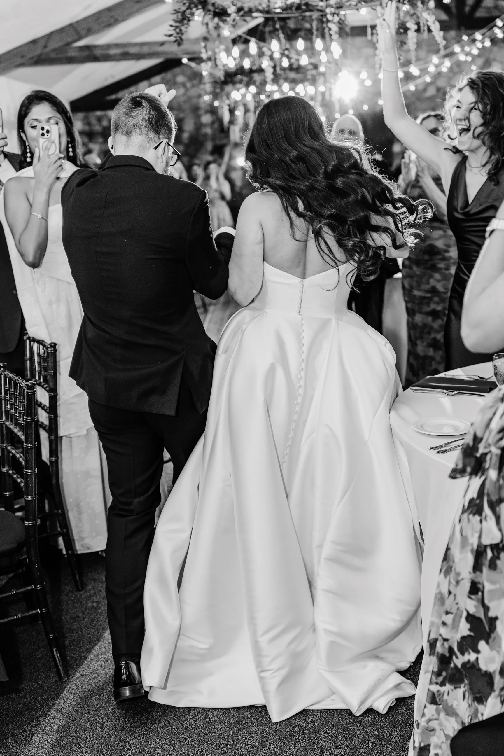 Black and white photograph of a wedding reception, showing a bride and groom dancing. The bride is wearing a strapless wedding gown with buttons down the back and voluminous skirt, and has long dark hair. The groom is in a dark suit. Guests are seen 