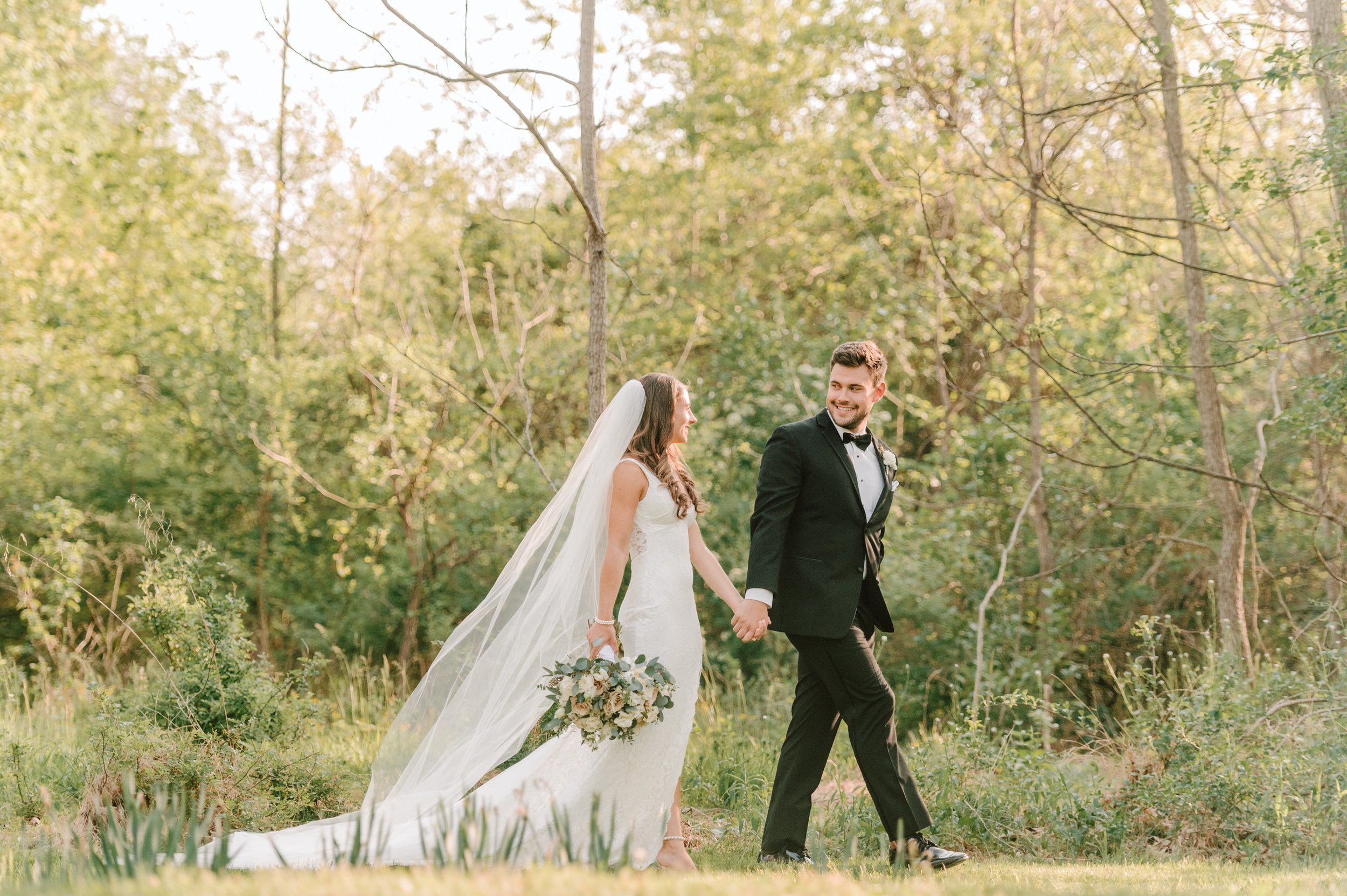 A happy bride and groom walking hand in hand through a green outdoor forested area during their wedding.