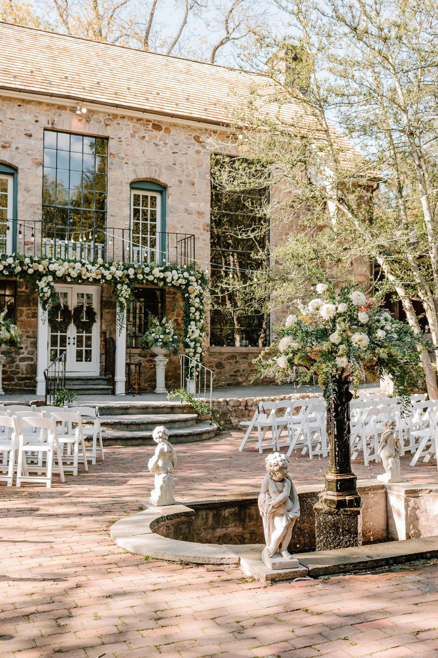 An outdoor setup for a wedding with white chairs arranged on a brick patio, a large floral arrangement on a pedestal, and a stone building decorated with white flowers in the background.