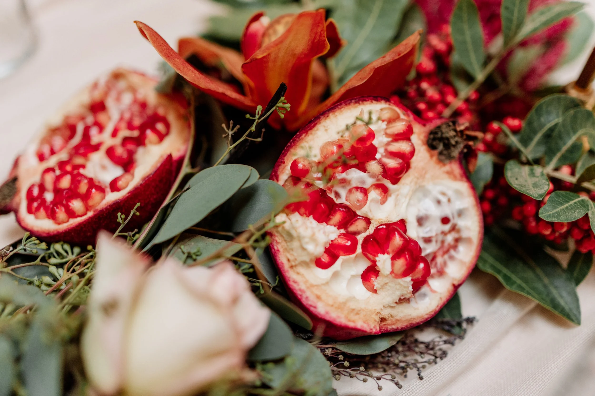 Close-up of a pomegranate cut open to reveal red seeds, surrounded by green leaves and floral accents.