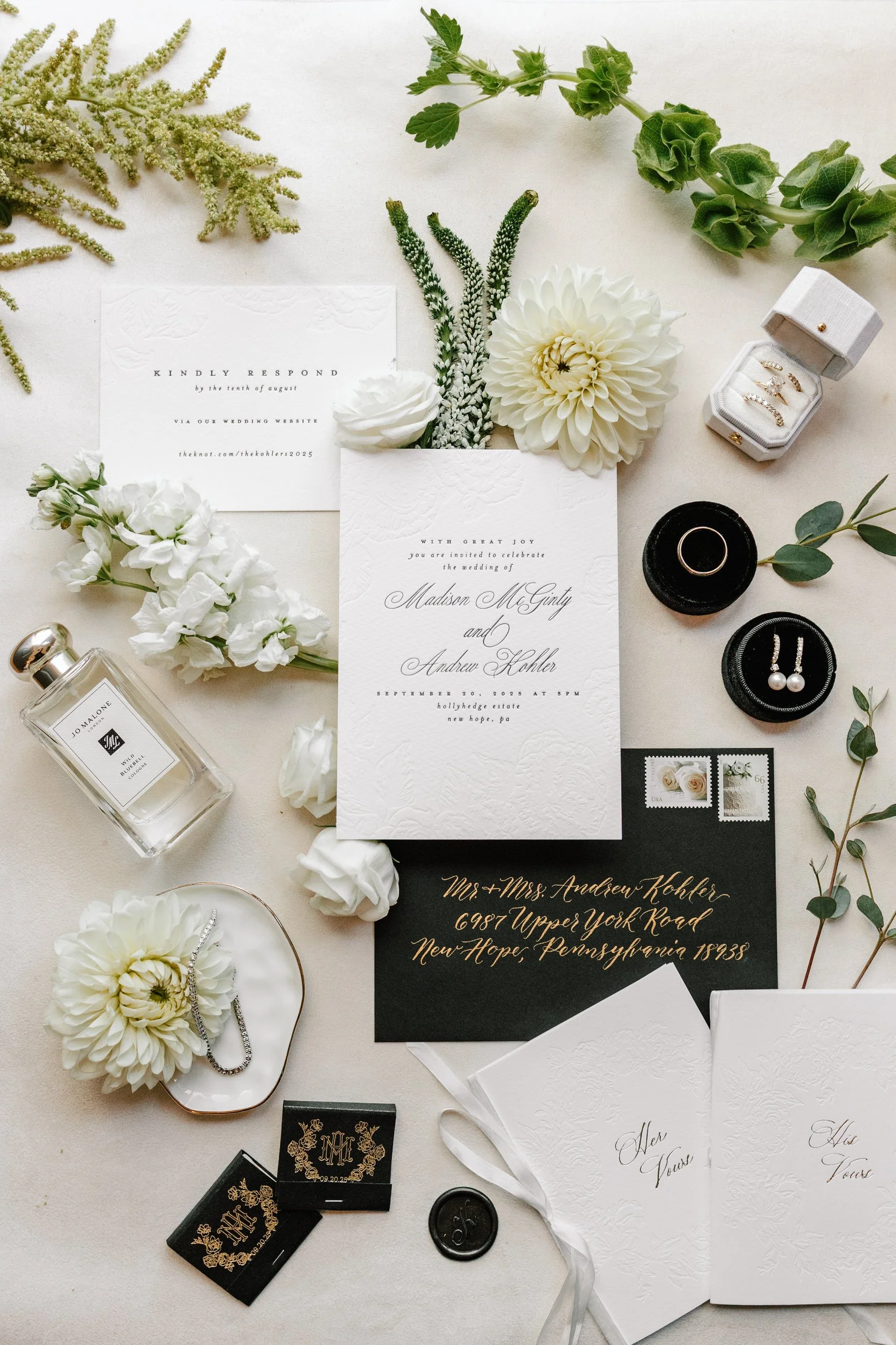 Wedding invitation suite with white and black stationery, floral arrangements, jewelry, perfume, and wedding accessories arranged on a white surface.