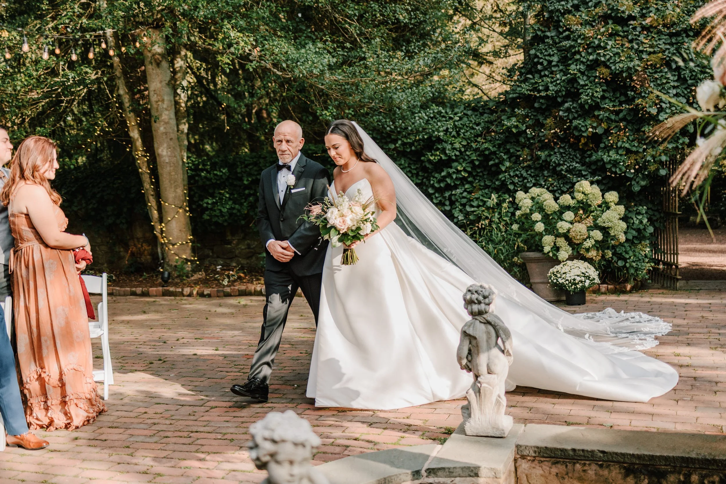 Bride walking with her father down an outdoor brick pathway at her wedding, surrounded by greenery and floral decorations.