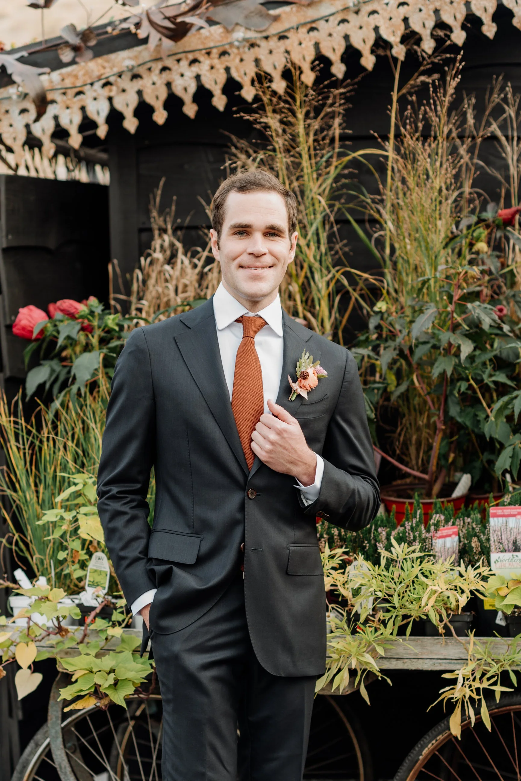 A man in a dark suit and orange tie stands outdoors in front of a garden with tall grasses and flowering plants, holding his lapel with his right hand and smiling.